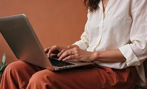 Person wearing white shirt and rust pants typing on a laptop with brown background.