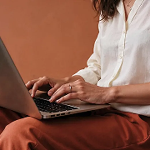 Person wearing white shirt and rust pants typing on a laptop with a brown background.