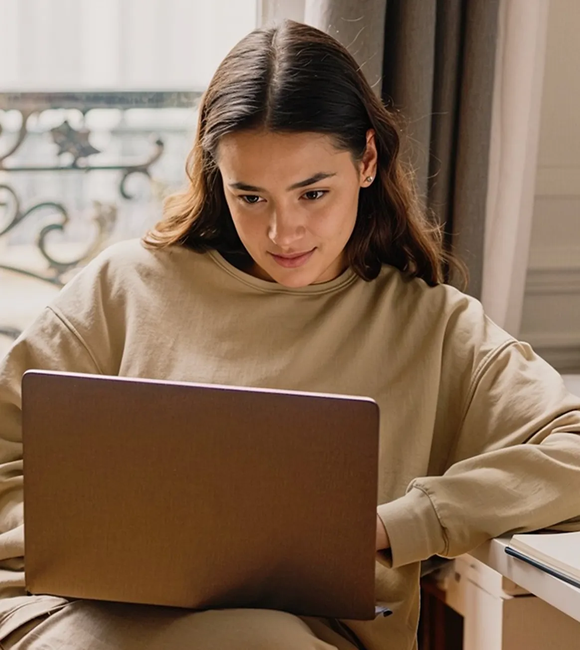Woman in tan sweater focused on laptop, sitting indoors with window and curtain in background.