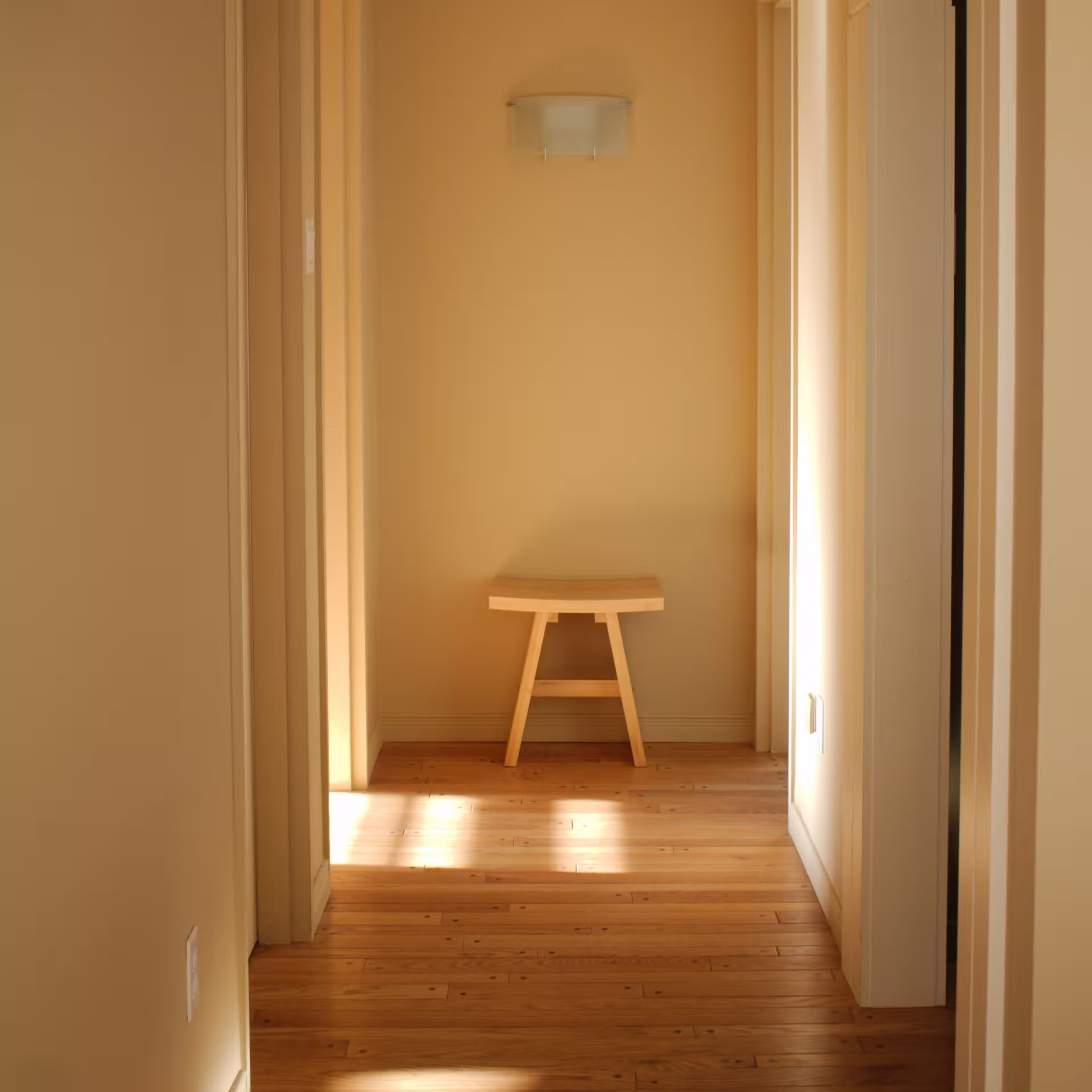 Simple wooden stool at the end of a warmly lit hallway with wood flooring and beige walls.