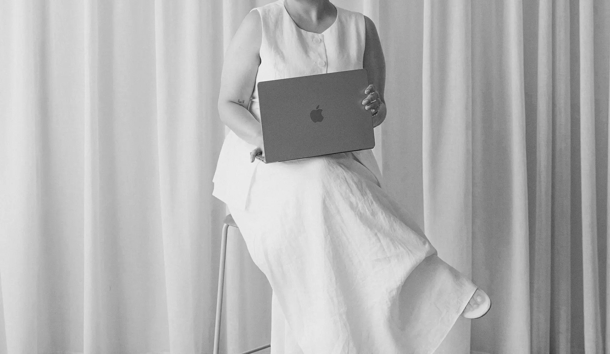 Person in a sleeveless white dress sitting with a laptop in front of a curtain backdrop.
