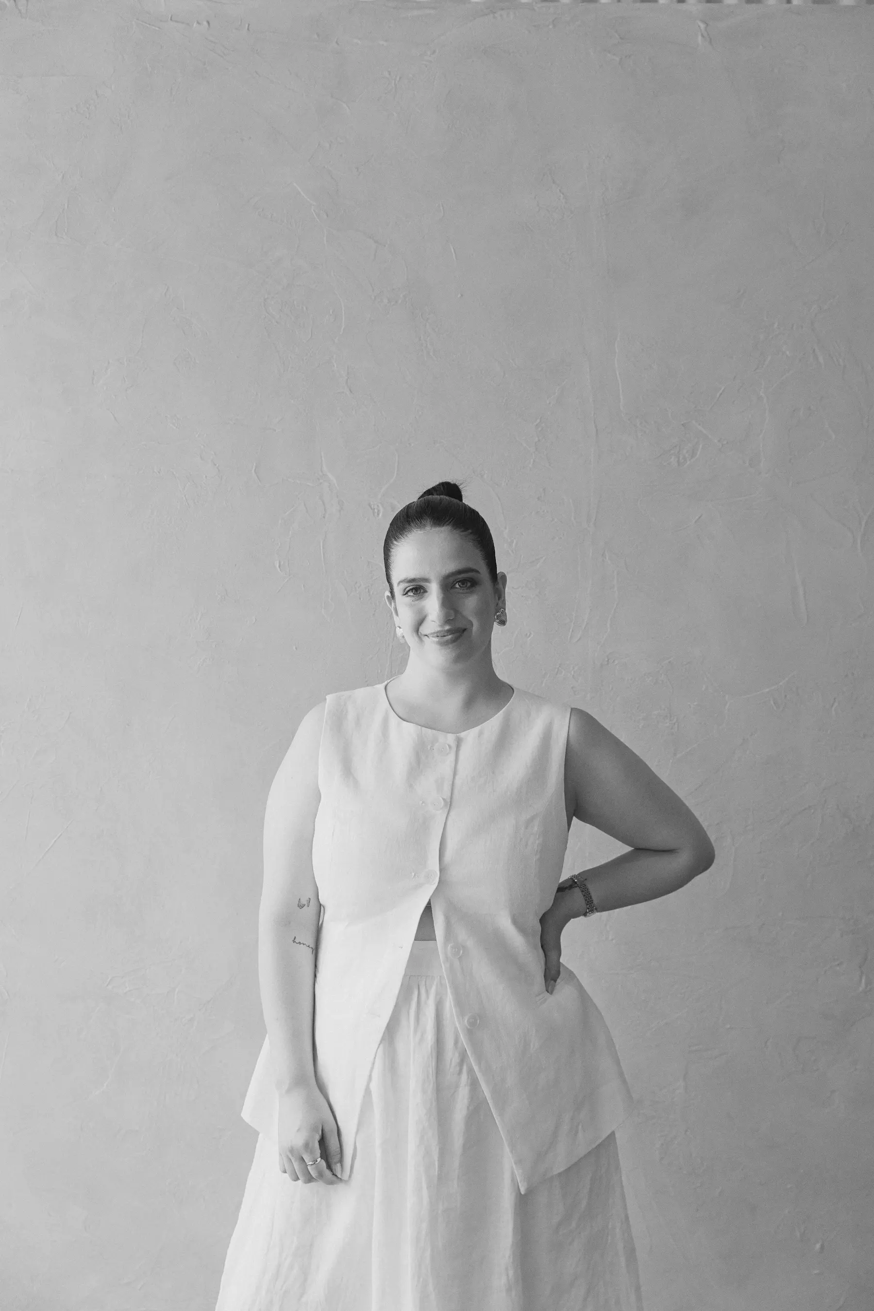 Smiling woman with hair in a bun wearing sleeveless buttoned top and skirt against textured wall.