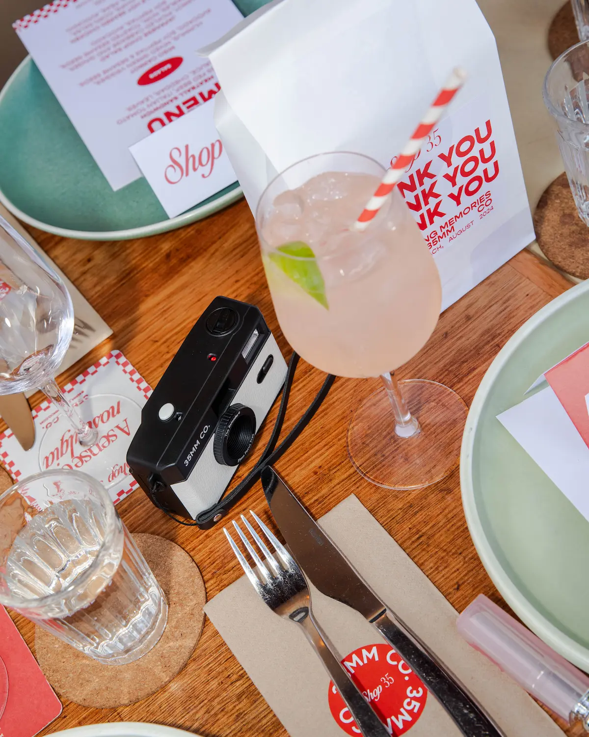Table setting with a pink drink, vintage camera, silverware on napkin, glass of water, and menus.