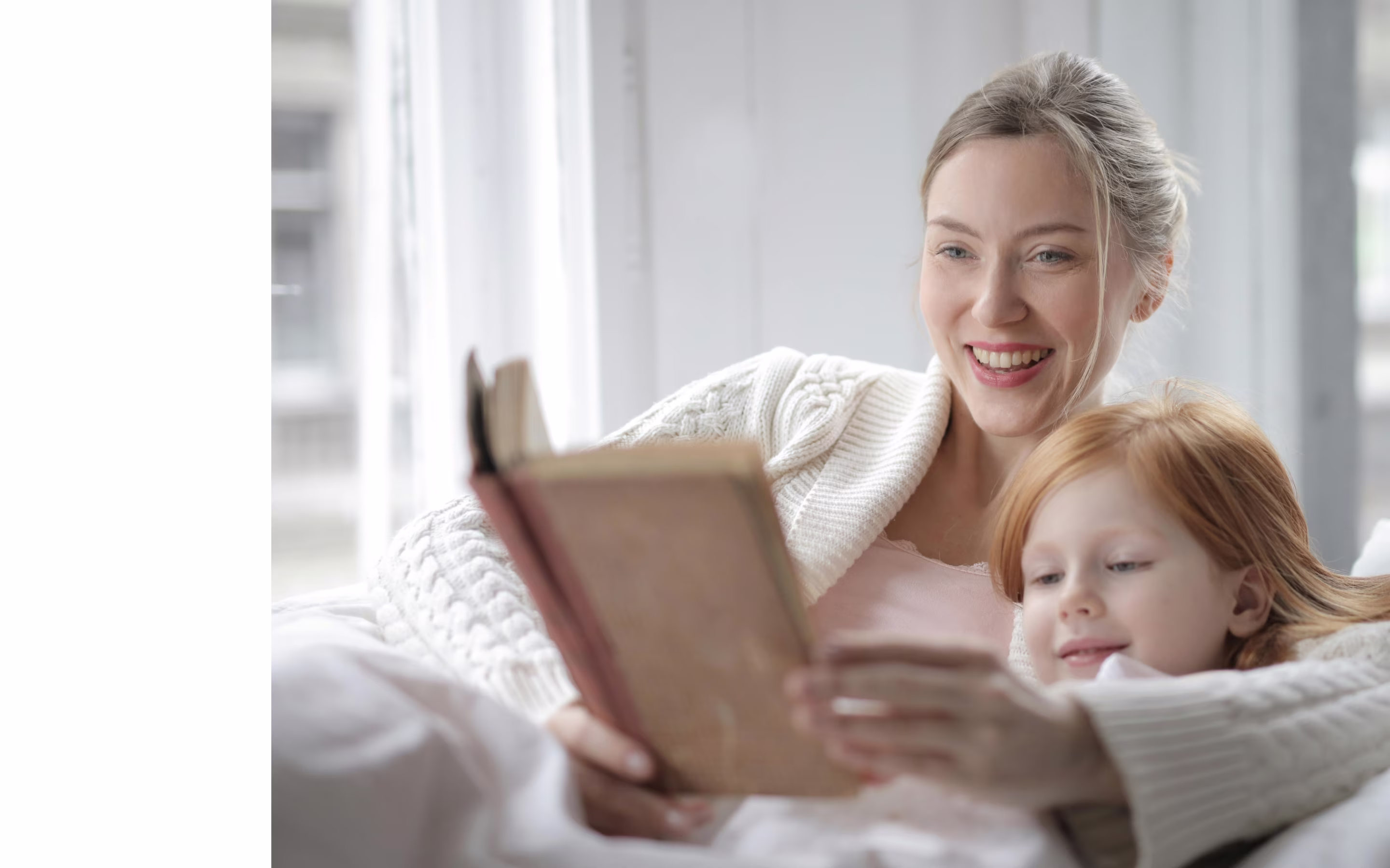 Smiling woman reading a book to a child on a couch by a bright window.