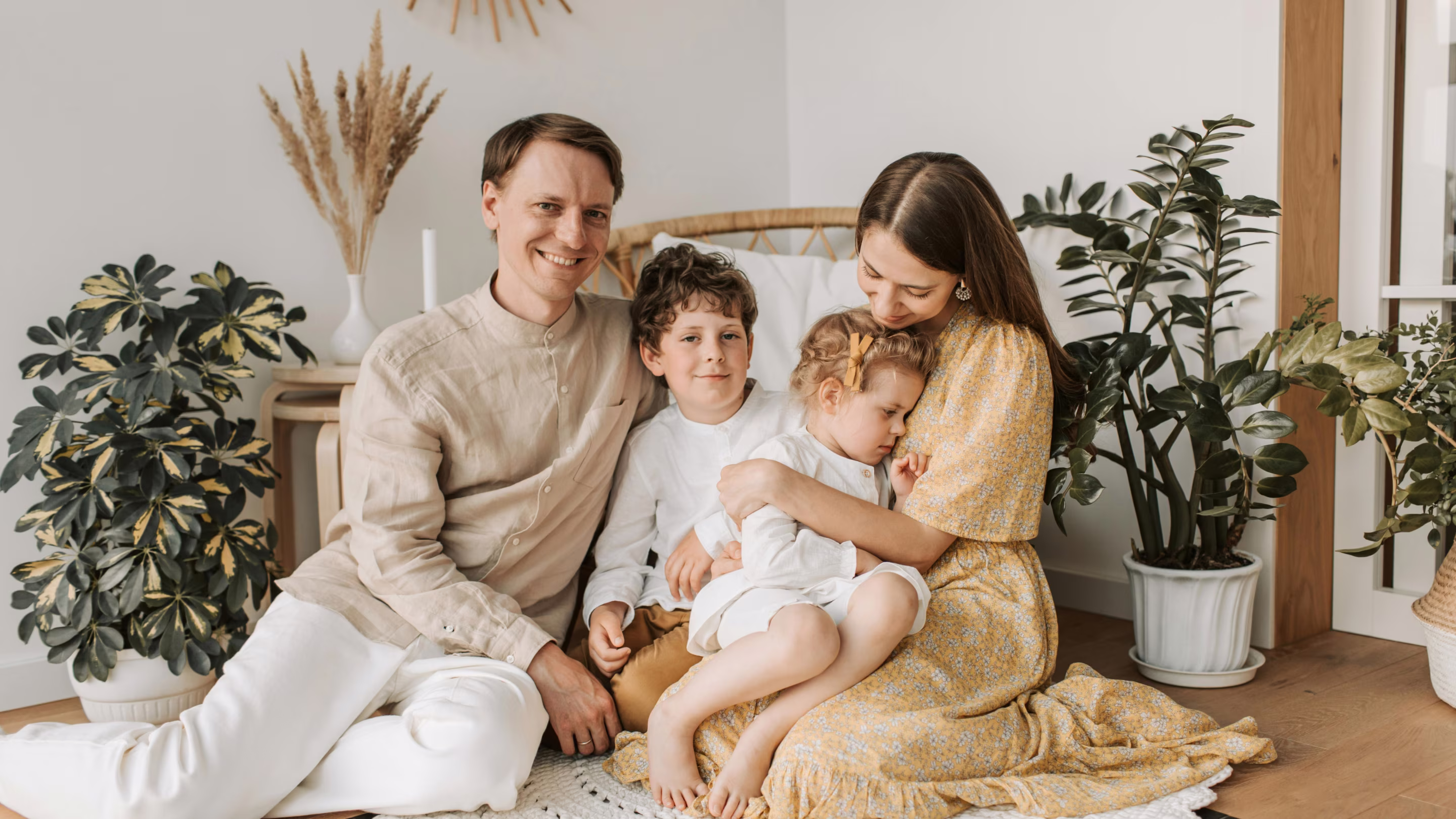 Smiling family sitting on the floor: man, boy, woman hugging a young girl in a cozy, plant-filled room.