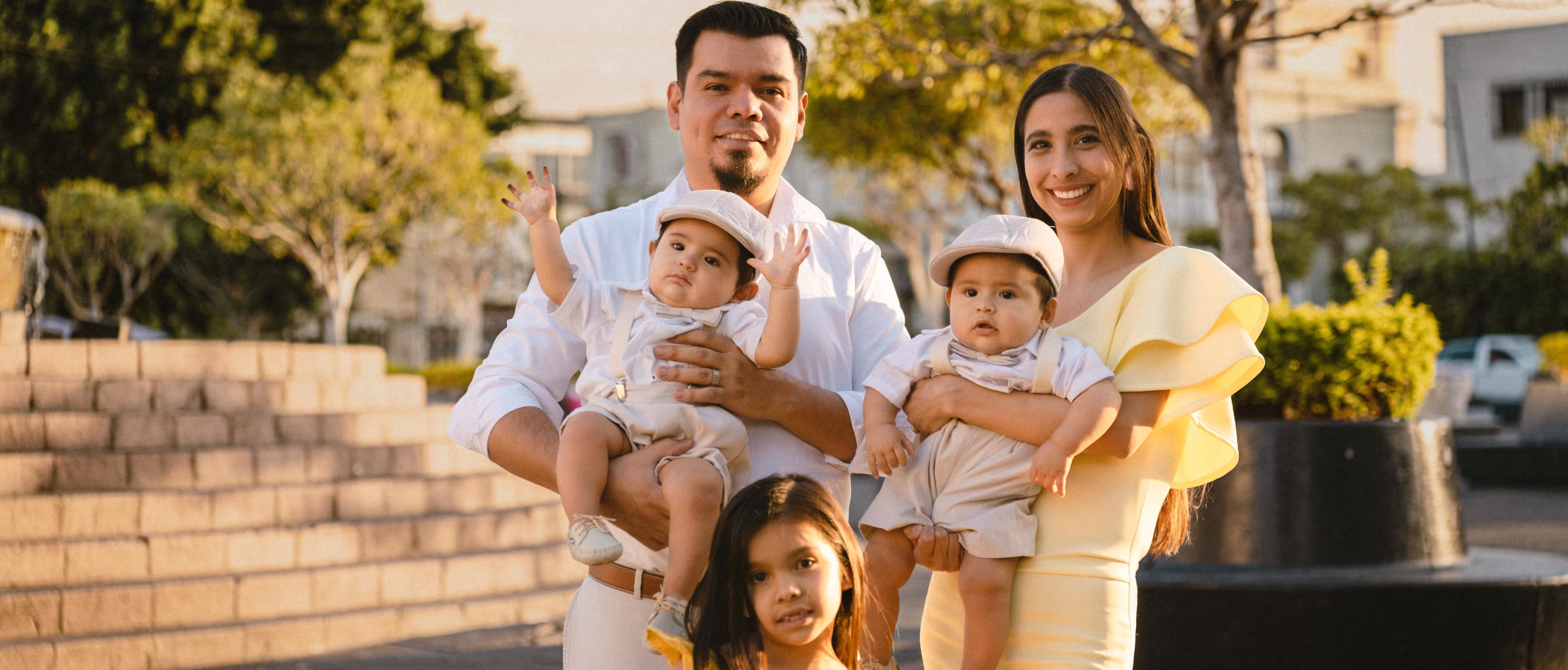 Smiling couple holding twin babies wearing hats and bowties with their young daughter outdoors.