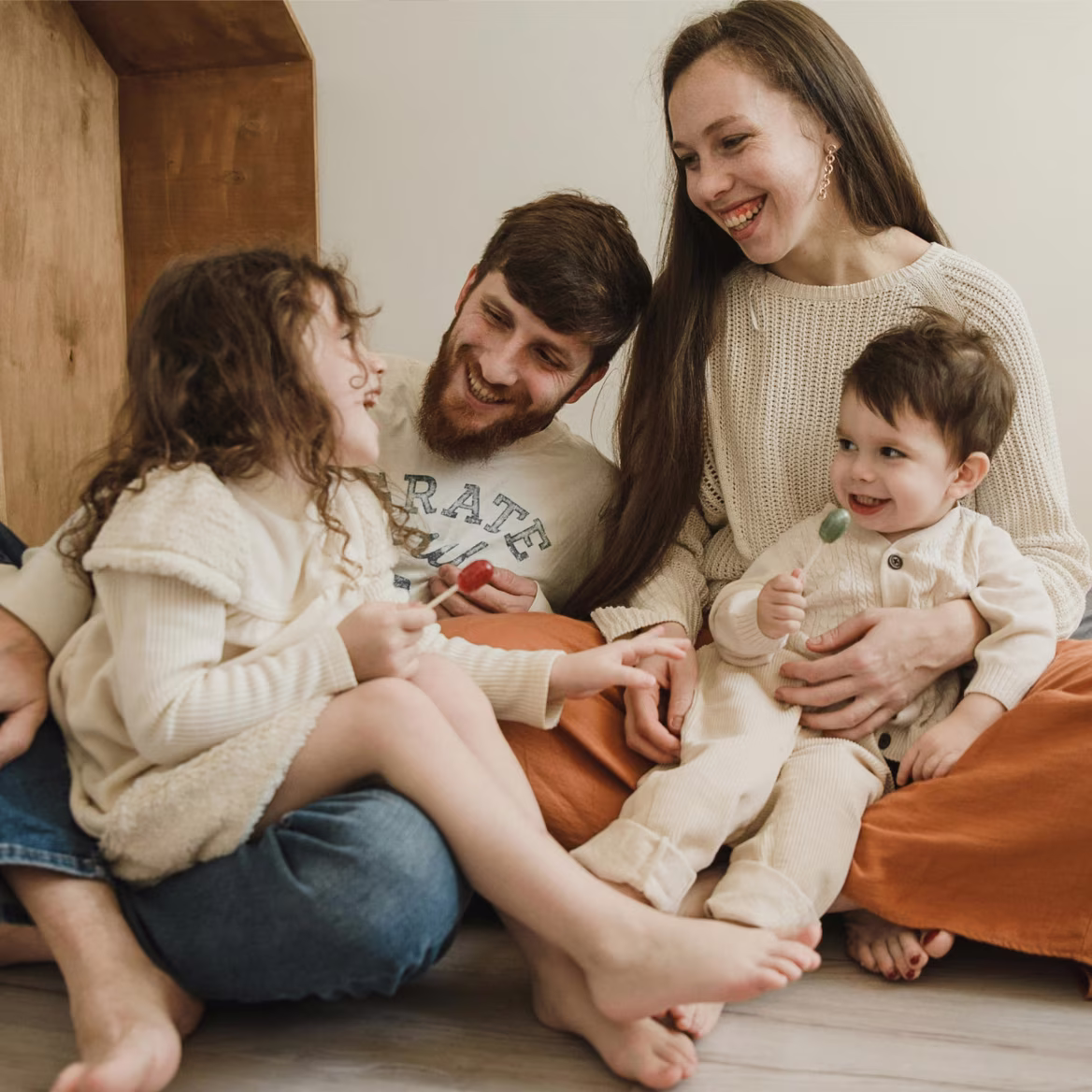 Smiling family sitting on floor: father, mother, daughter, and toddler holding lollipops and laughing.