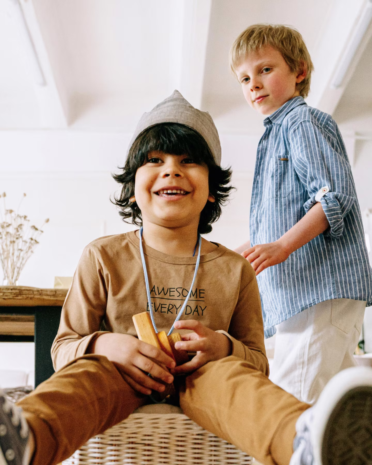 Two young boys indoors, one sitting and smiling with wooden blocks, the other standing nearby.