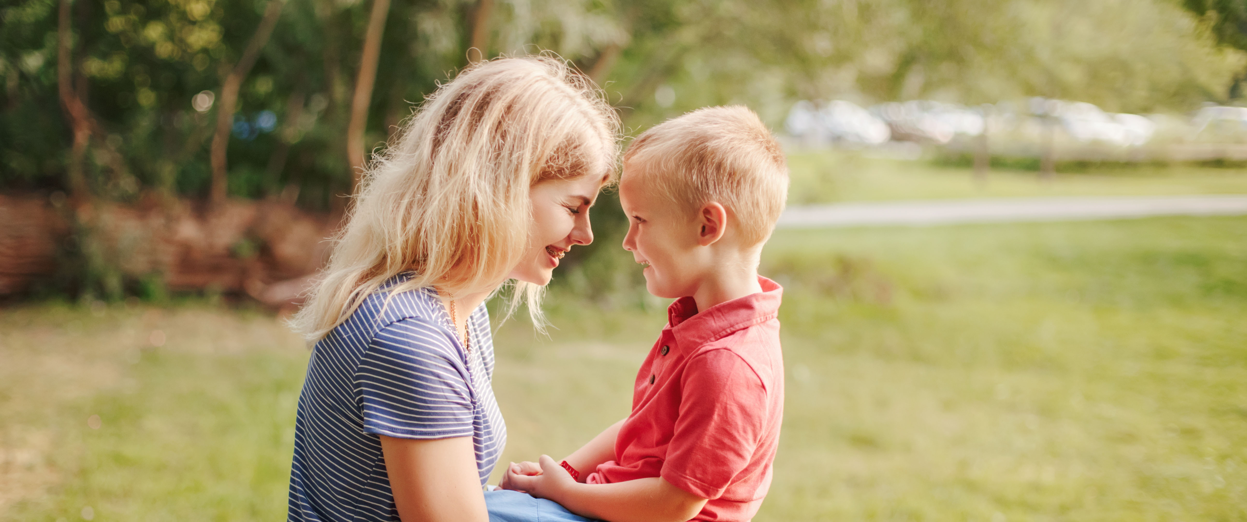 An adult and a young child sit facing each other outdoors, foreheads almost touching, both smiling.