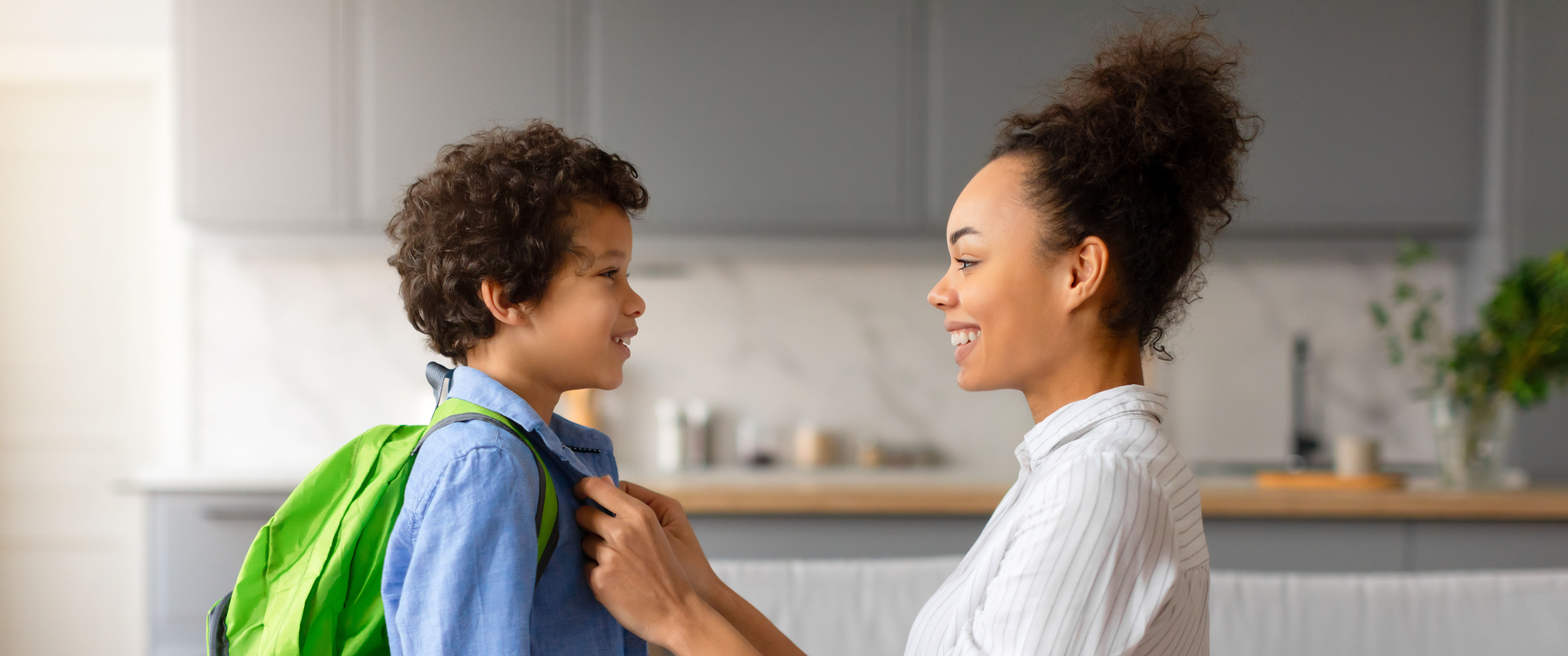 An adult adjusts the collar of a child wearing a backpack, the two looking at each other.