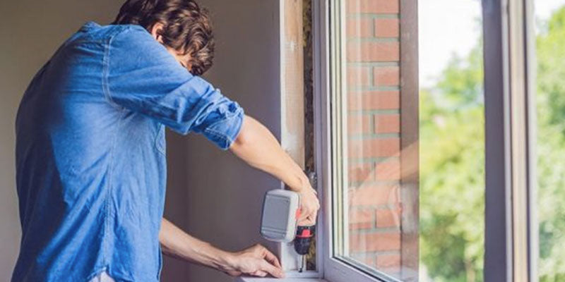 Person installing a window with a drill, wearing a blue shirt.