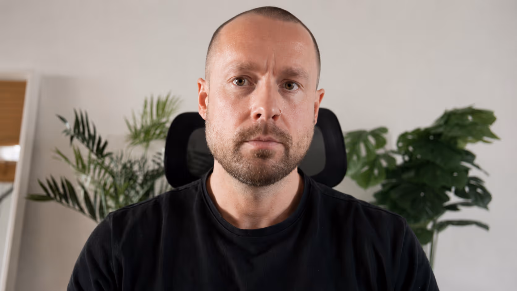 Man with short beard and shaved head sitting in front of green plants in a black chair.