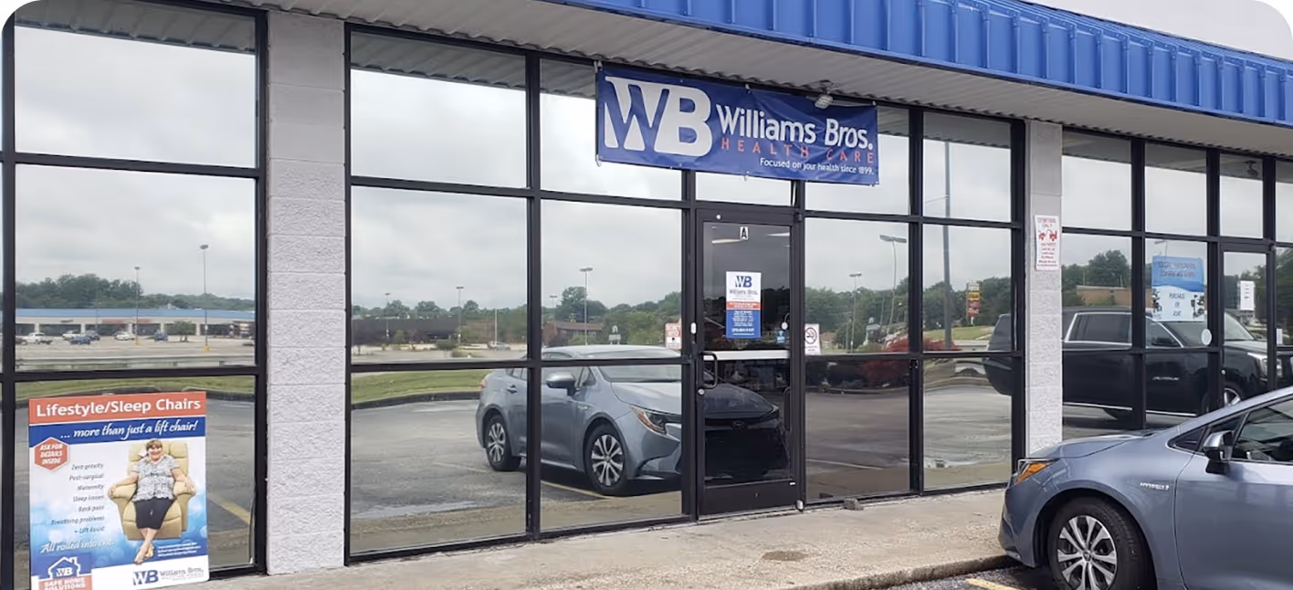 Glass storefront of Williams Bros. Healthcare with blue sign and parked cars in reflections and front.