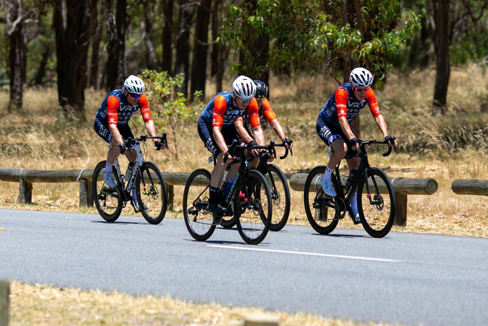Four cyclists in matching blue and orange gear riding road bikes on a paved path in a wooded area.