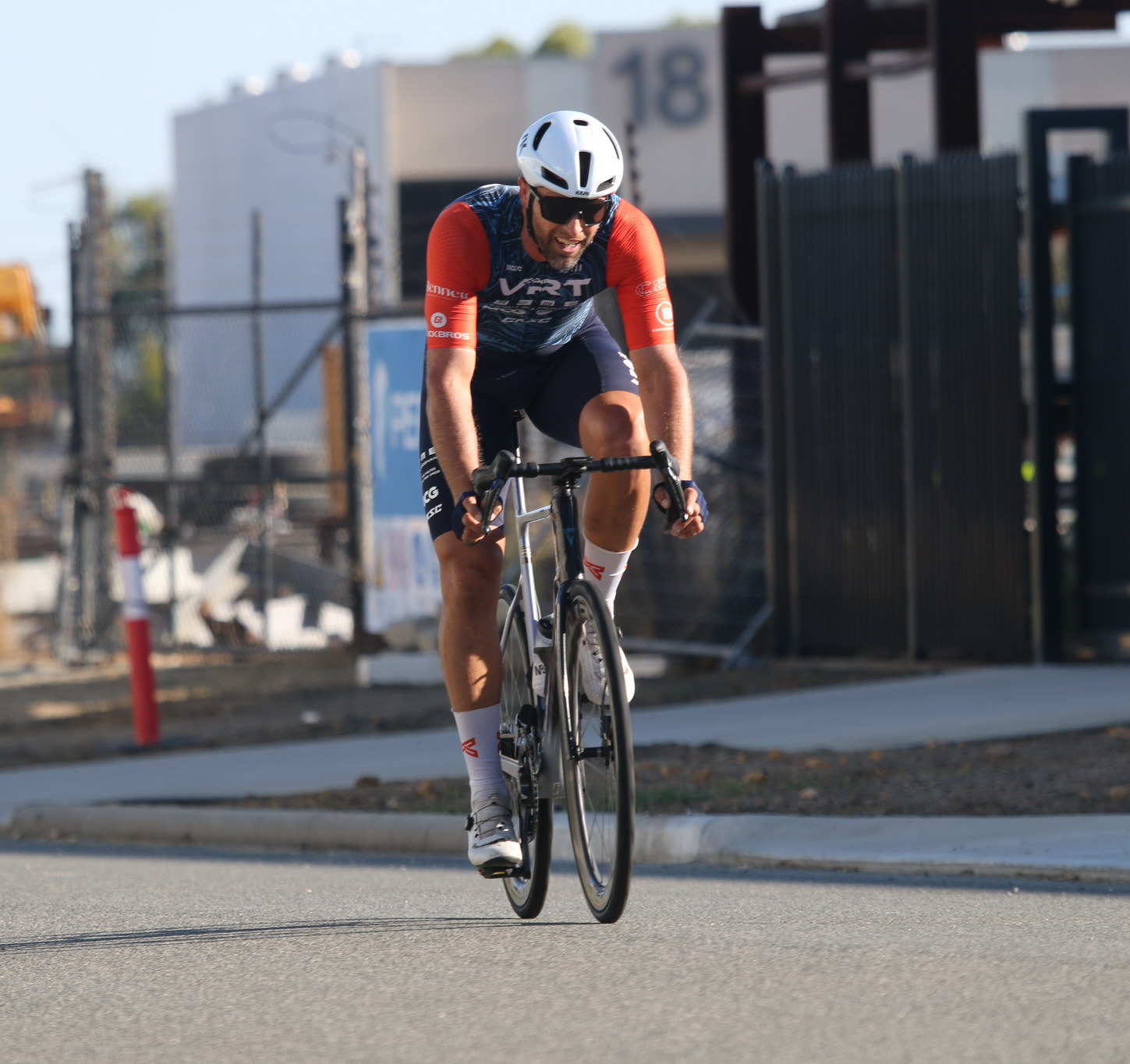 Cyclist wearing a helmet and sunglasses rides a road bike on a paved street near a fenced area.
