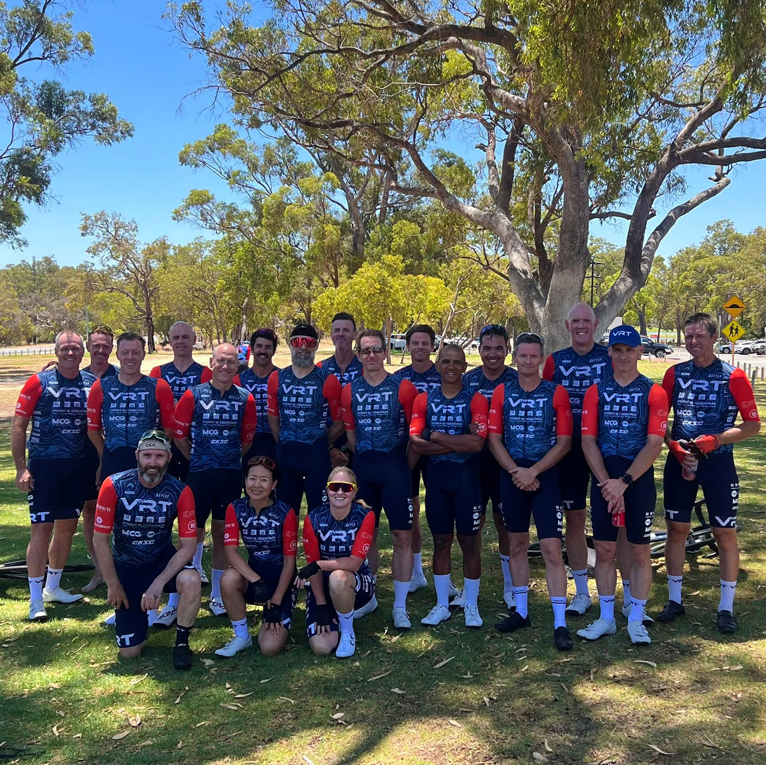 Cycling team in blue and red VRT jerseys posing outdoors on grass under trees on a sunny day.