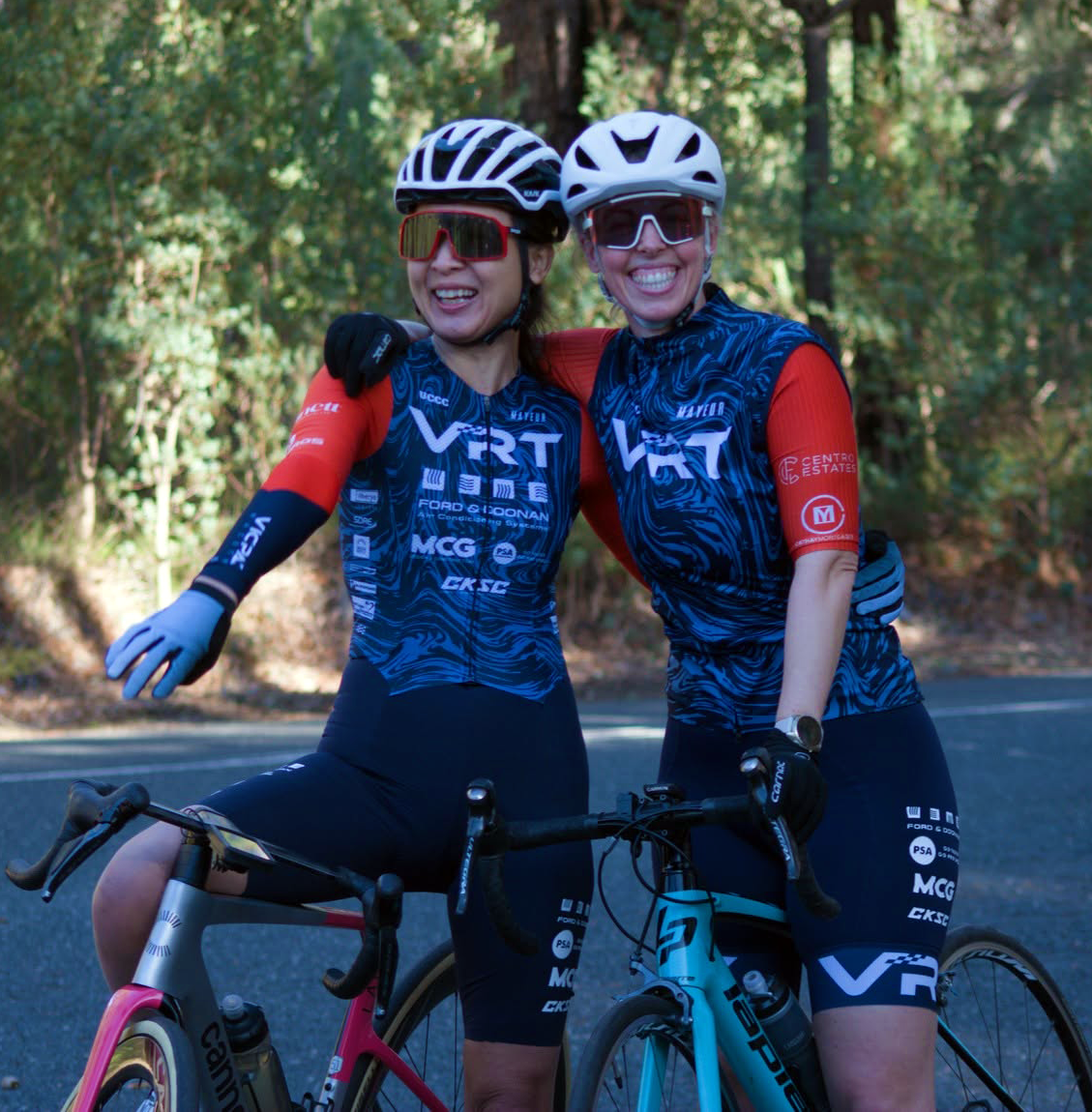 Two smiling cyclists in matching blue and orange VRT team jerseys with helmets and sunglasses, outdoors.