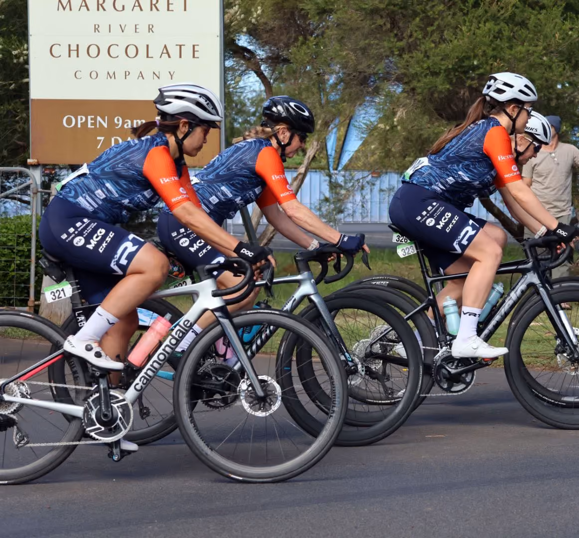 Three cyclists in matching blue and orange jerseys ride racing bikes near a Margaret River Chocolate Company sign.