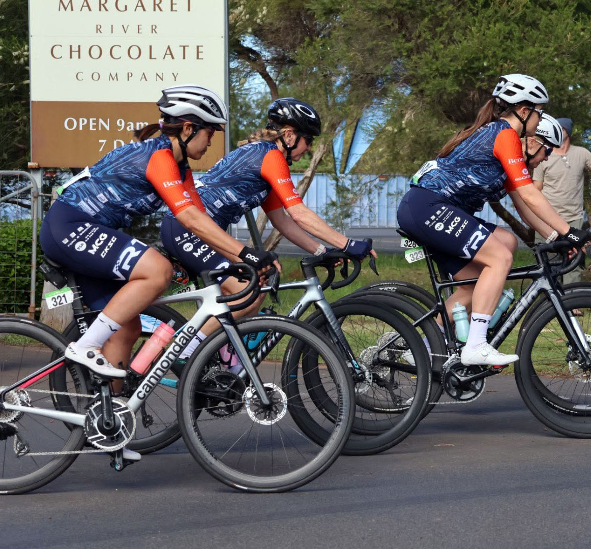 Three cyclists in matching blue and orange jerseys ride racing bikes near a Margaret River Chocolate Company sign.