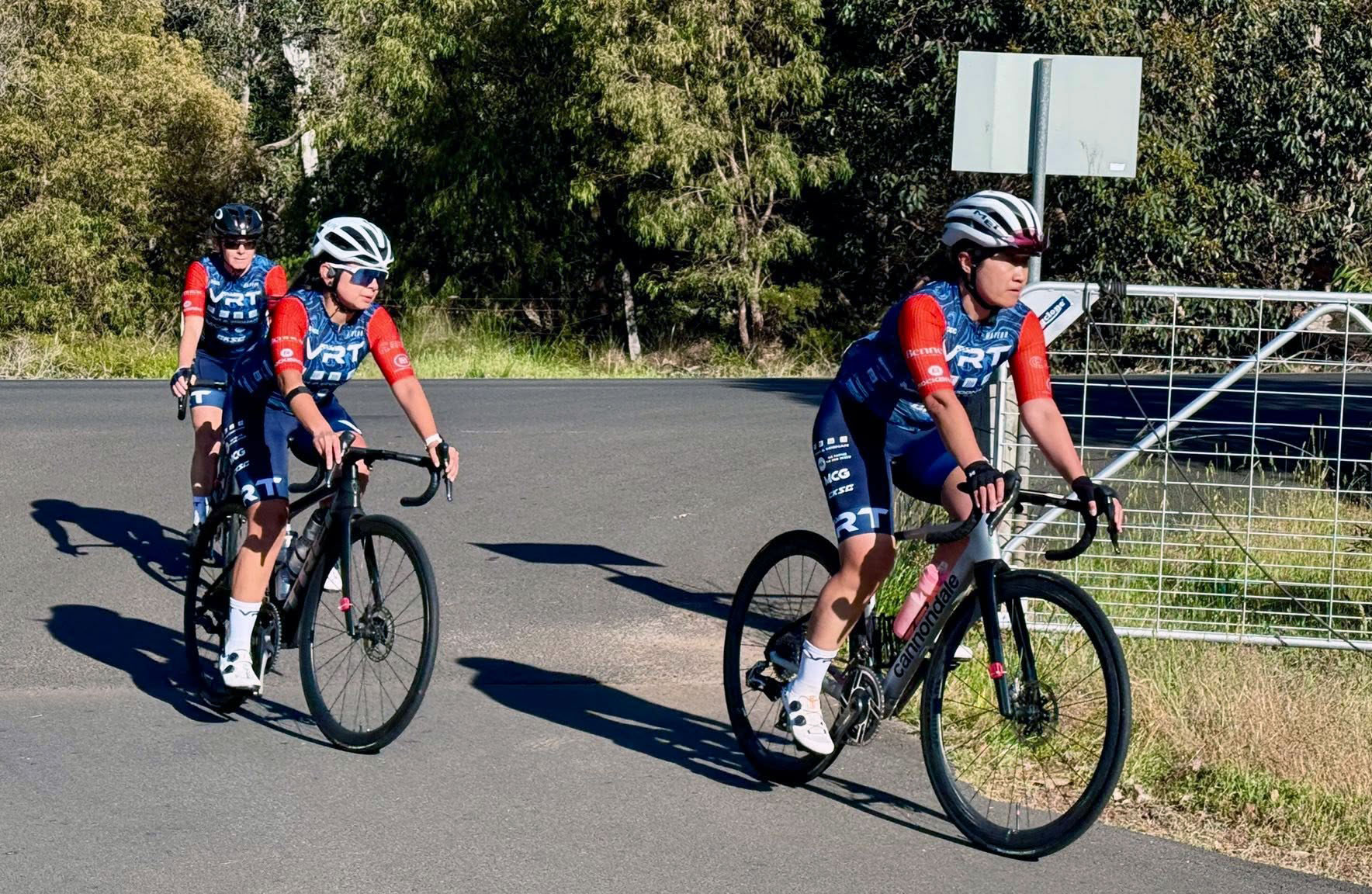 Three cyclists in matching blue and red gear riding on a road surrounded by greenery.