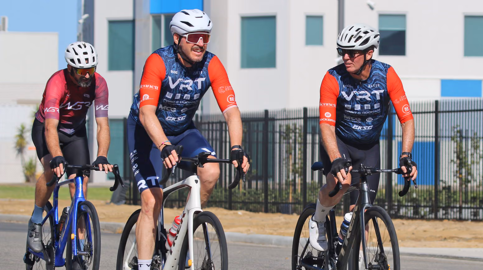 Three cyclists wearing helmets and jerseys riding road bikes on a sunny urban street.