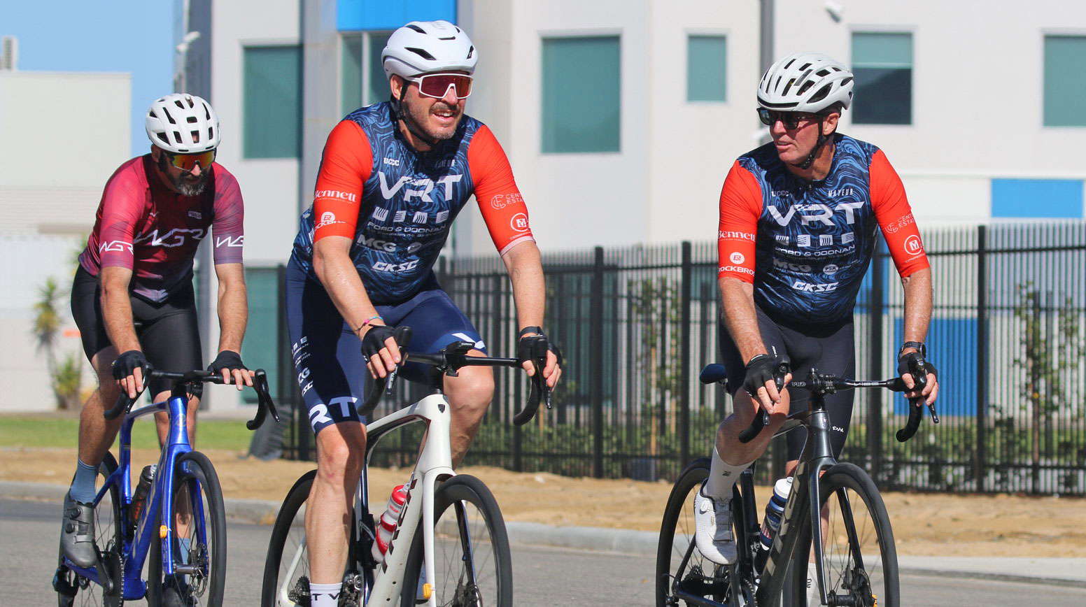 Three cyclists wearing helmets and jerseys riding road bikes on a street in front of a building.