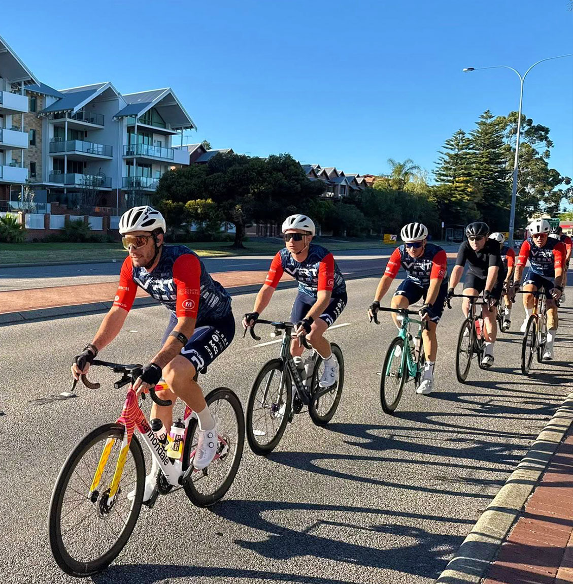 Group of cyclists in matching jerseys training on a sunny street near residential buildings.