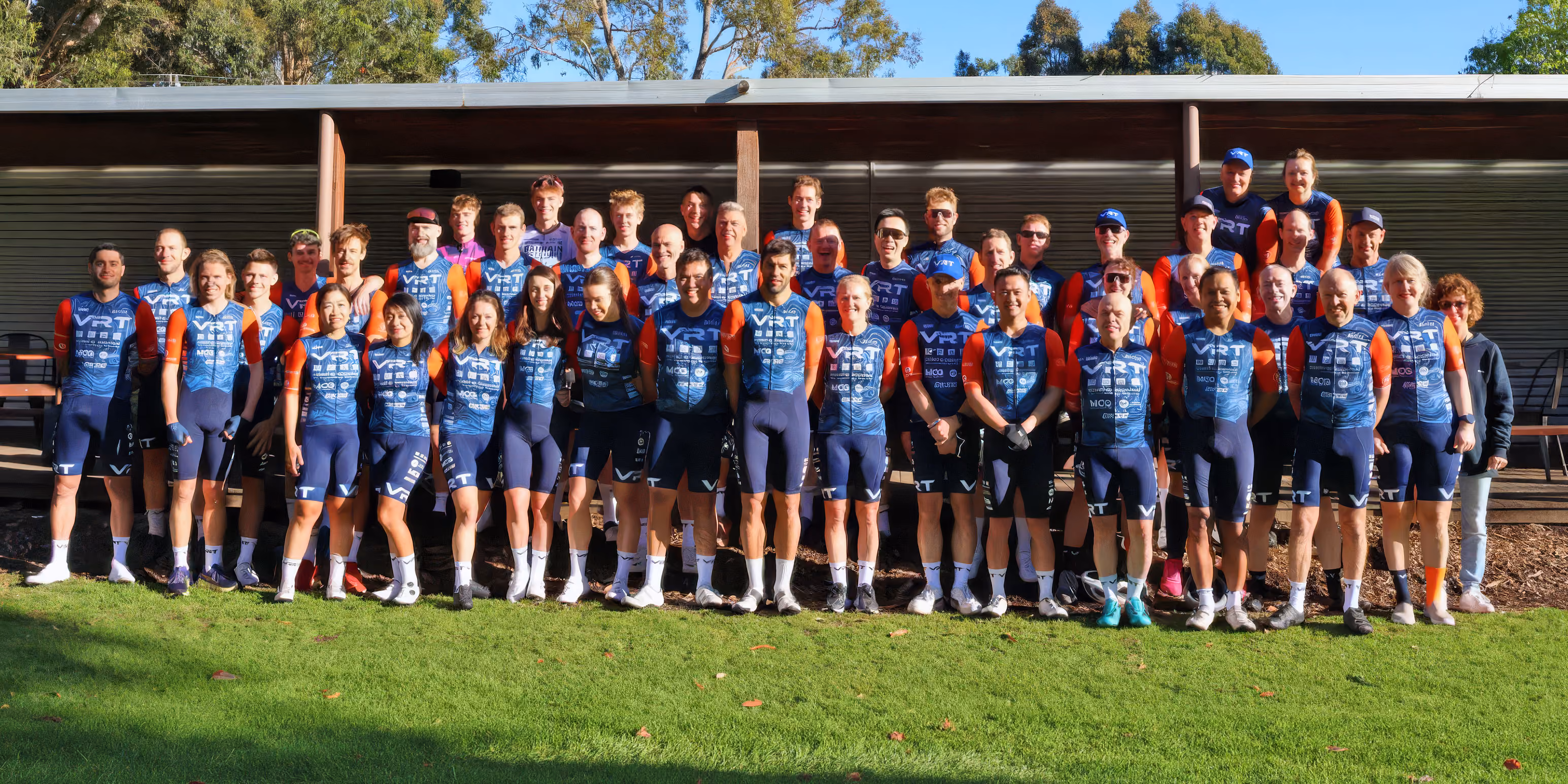 Three cyclists in matching VRT race team uniforms posing outdoors under blue sky by a sponsor banner.