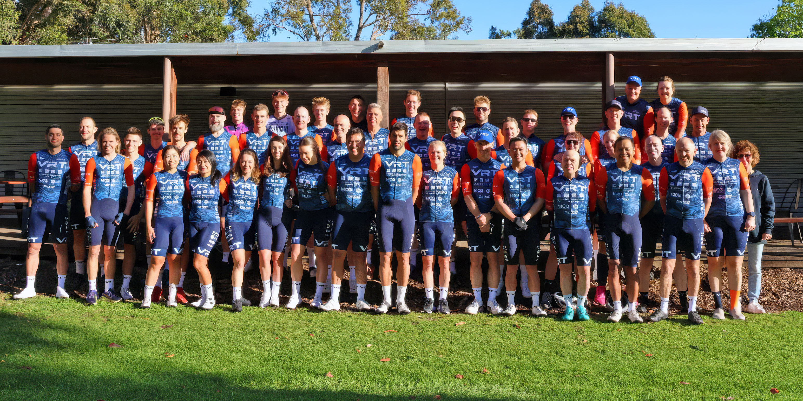 Three cyclists in matching VRT race team uniforms posing outdoors under blue sky by a sponsor banner.