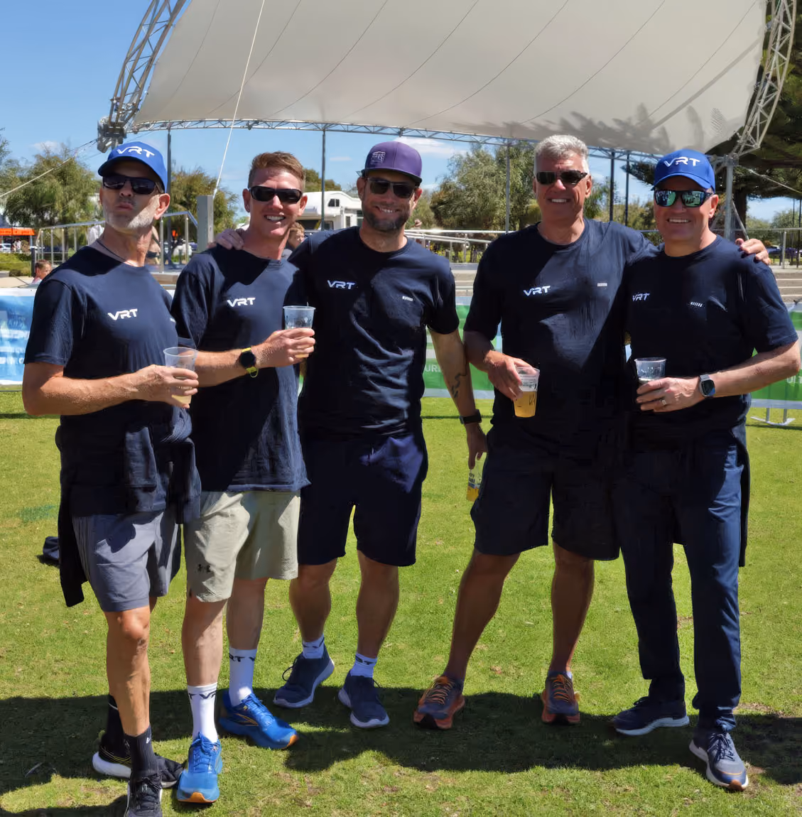 Five men wearing matching VRT shirts and sunglasses standing on grass holding drinks under a white canopy.
