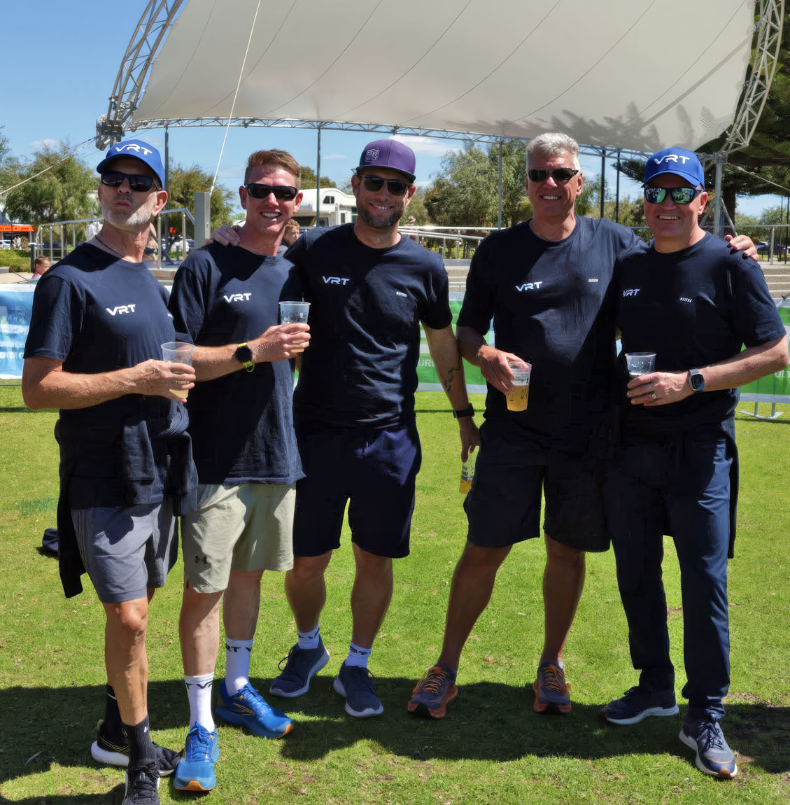 Five men wearing matching VRT shirts and sunglasses standing on grass holding drinks under a white canopy.