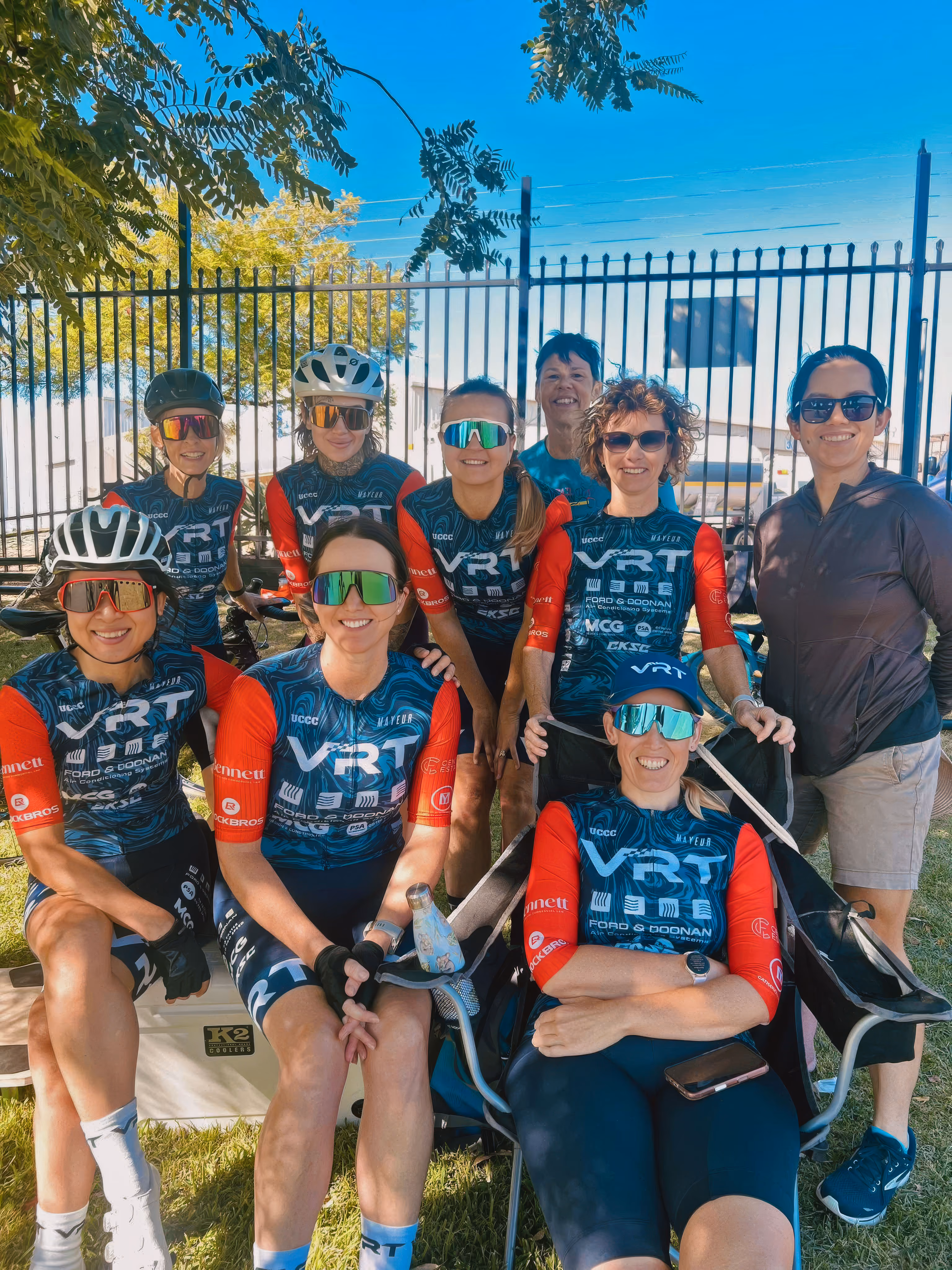 Group of eight women cyclists in team uniforms posing outdoors with smiles under tree shade.