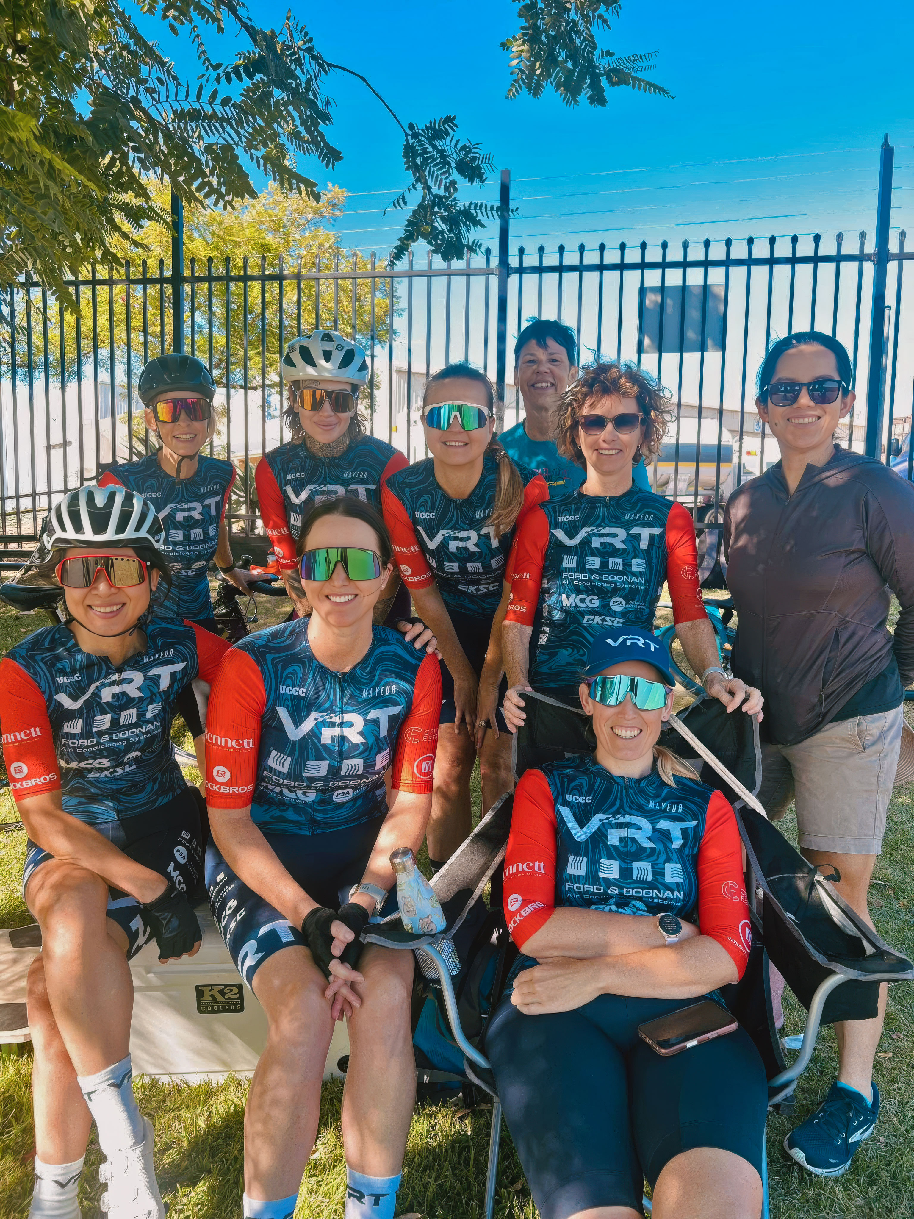 Group of eight women cyclists in team uniforms posing outdoors with smiles under tree shade.