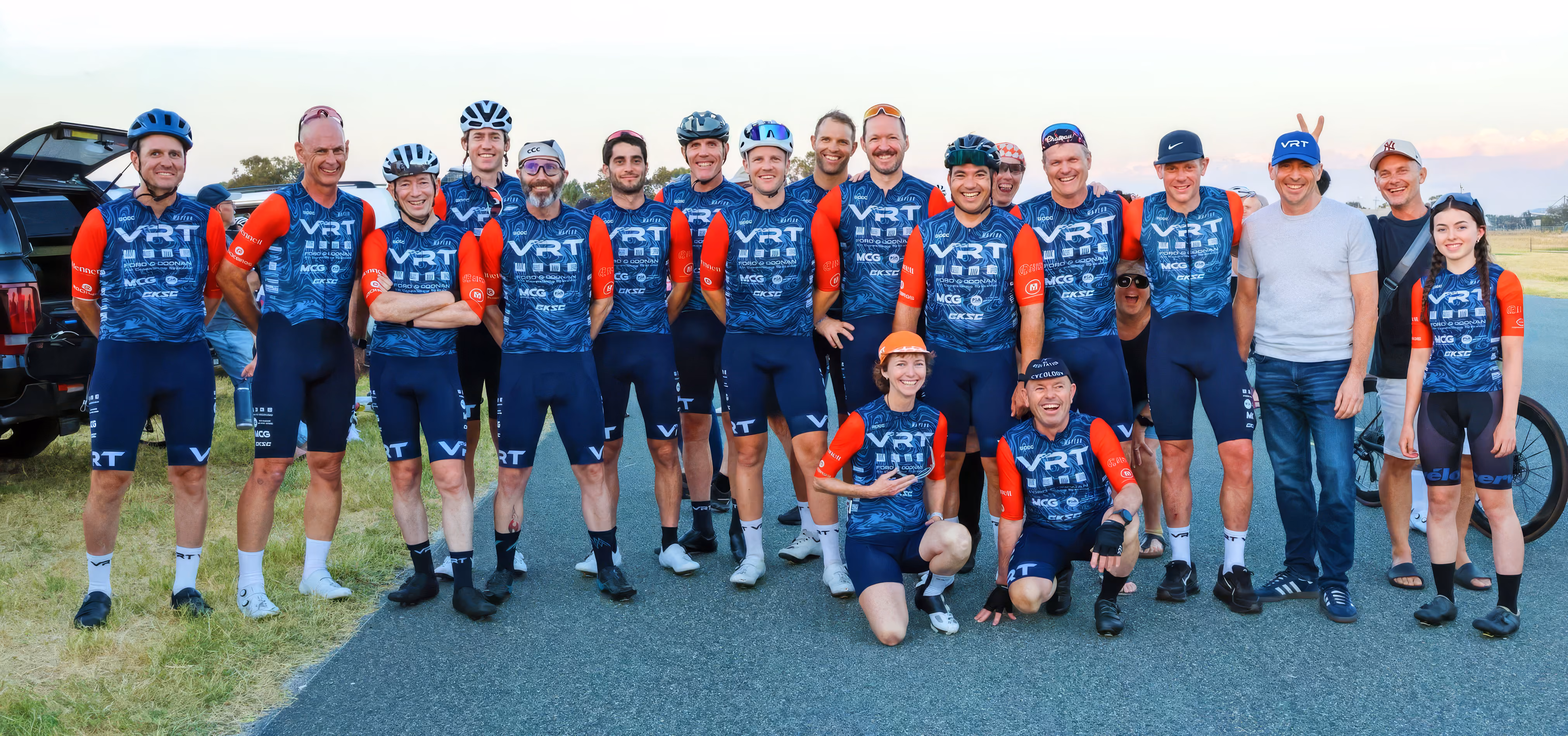 Group of cyclists in blue and red VRT racing jerseys posing outdoors on road and grass.