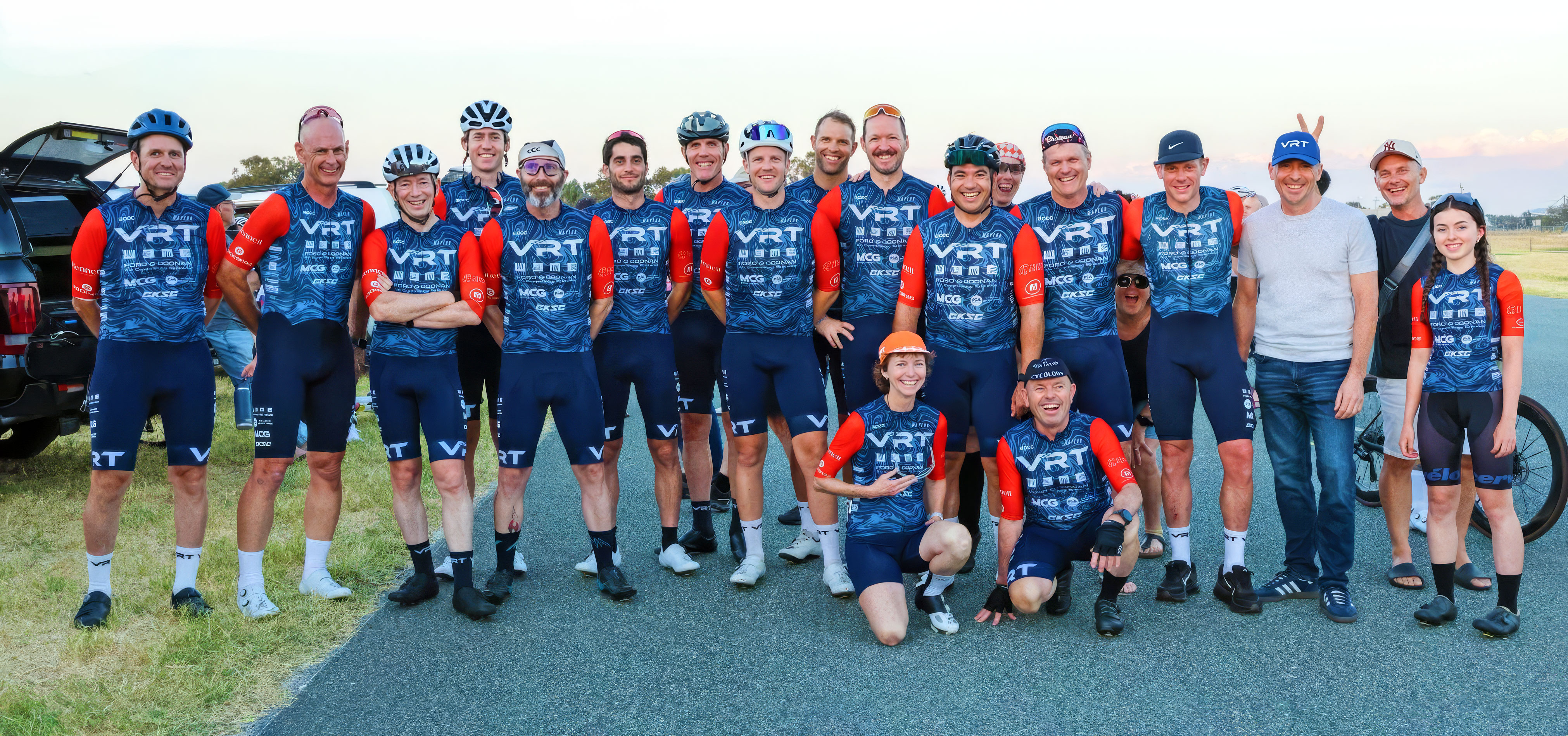 Group of cyclists in blue and red VRT racing jerseys posing outdoors on road and grass.