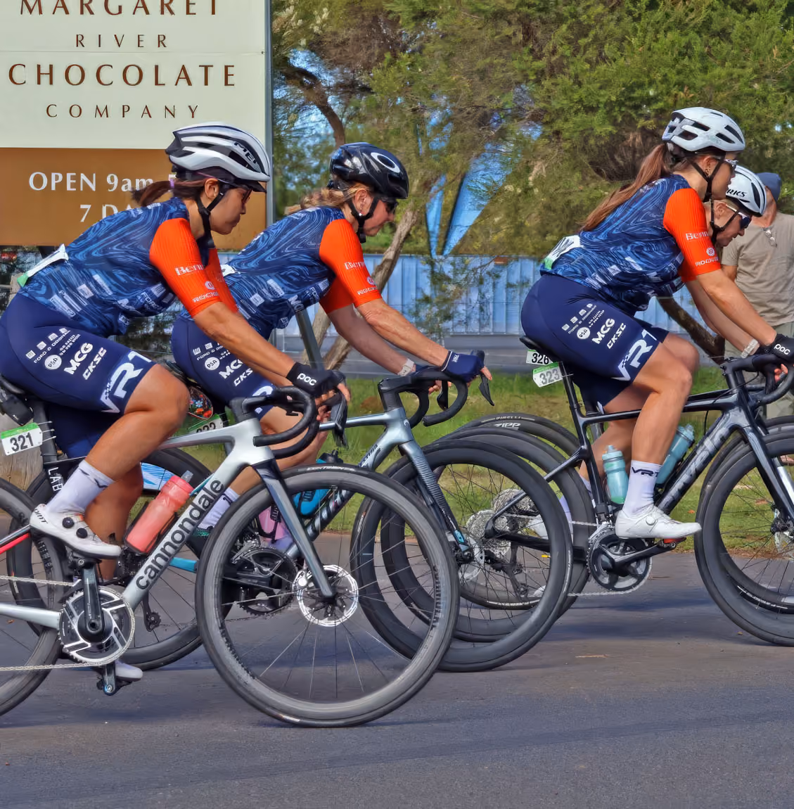 Three female cyclists in matching blue and orange gear race past a Margaret River Chocolate Company sign.