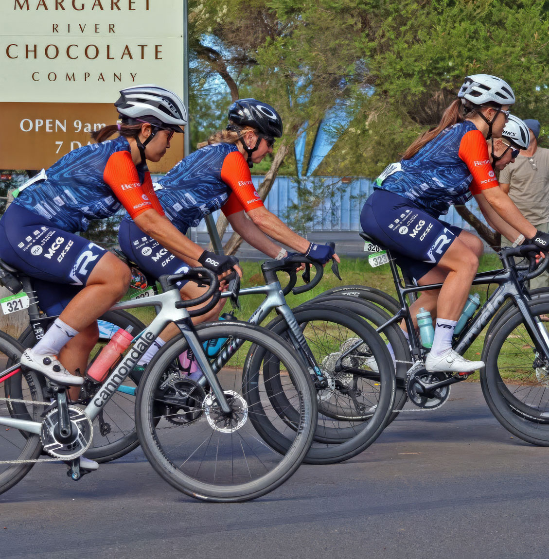 Three female cyclists in matching blue and orange gear race past a Margaret River Chocolate Company sign.