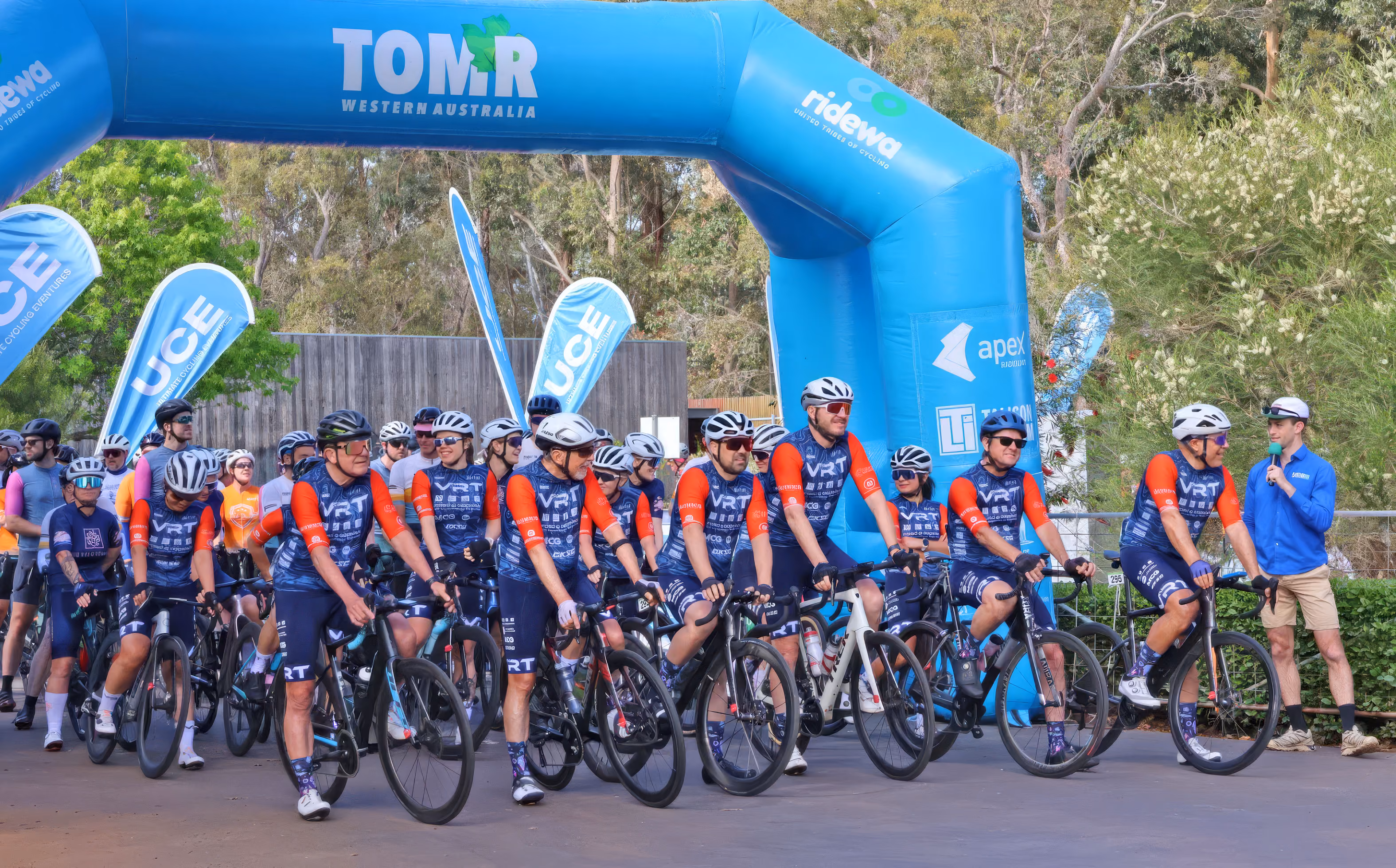 Group of cyclists in blue and orange gear gathered at start line under a blue TOM:R Western Australia arch.