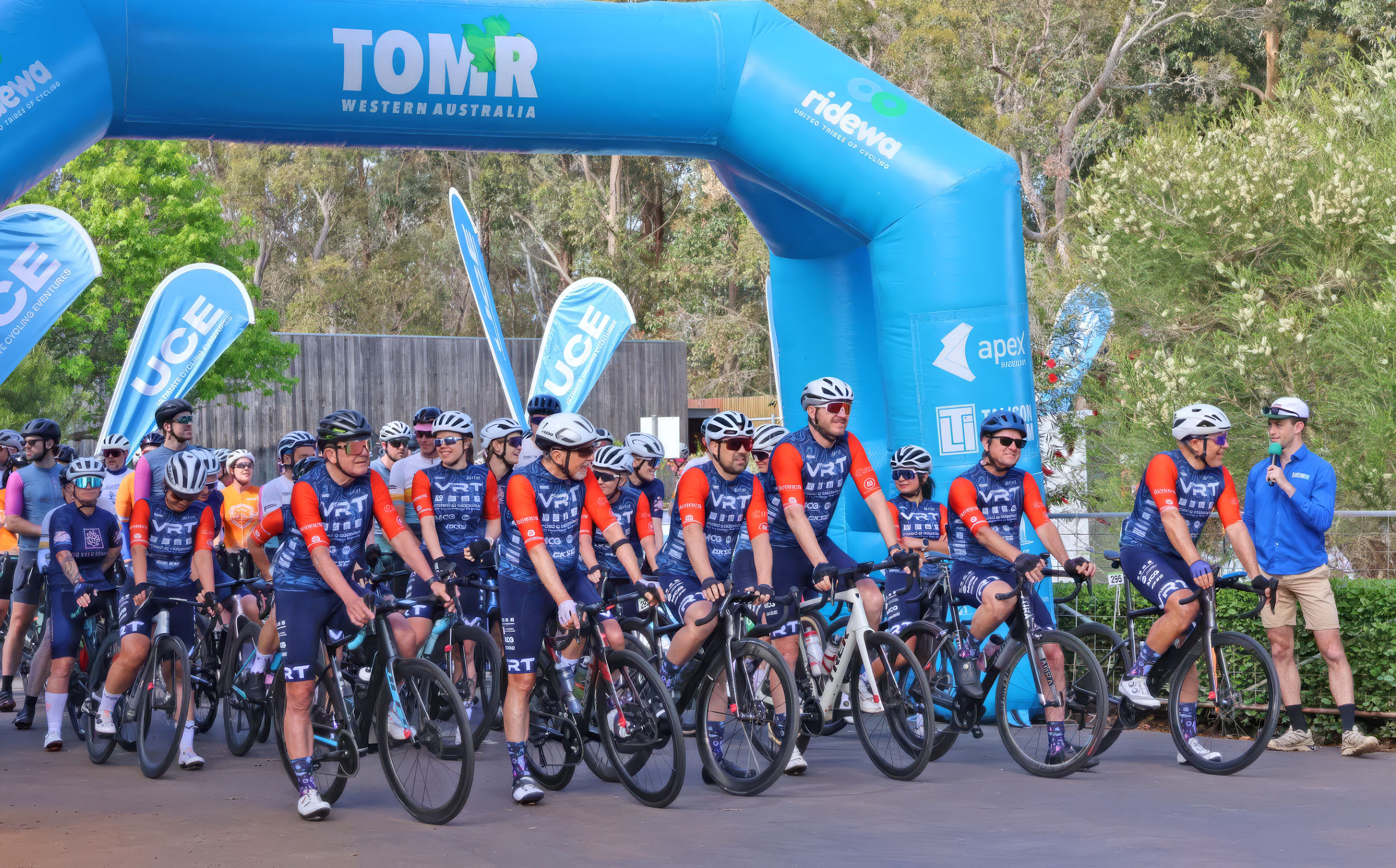 Group of cyclists in blue and orange gear gathered at start line under a blue TOM:R Western Australia arch.