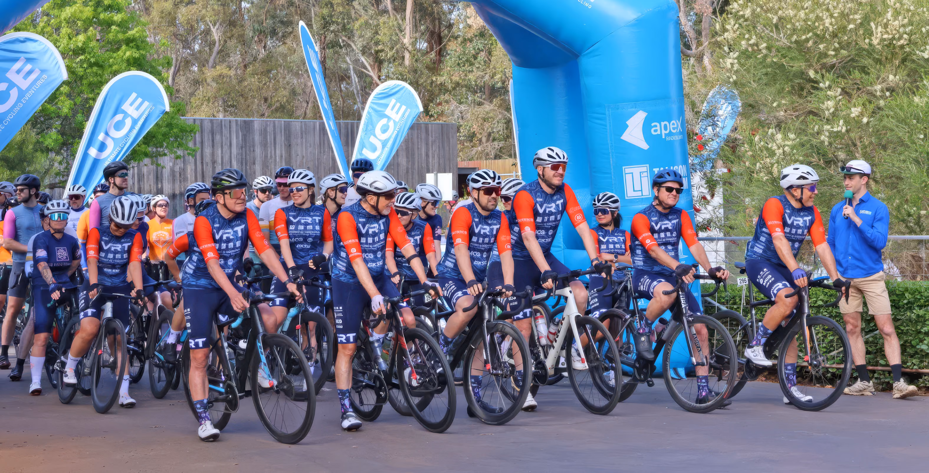 Cycling team in matching blue and orange jerseys on bikes at event start with announcer speaking nearby.