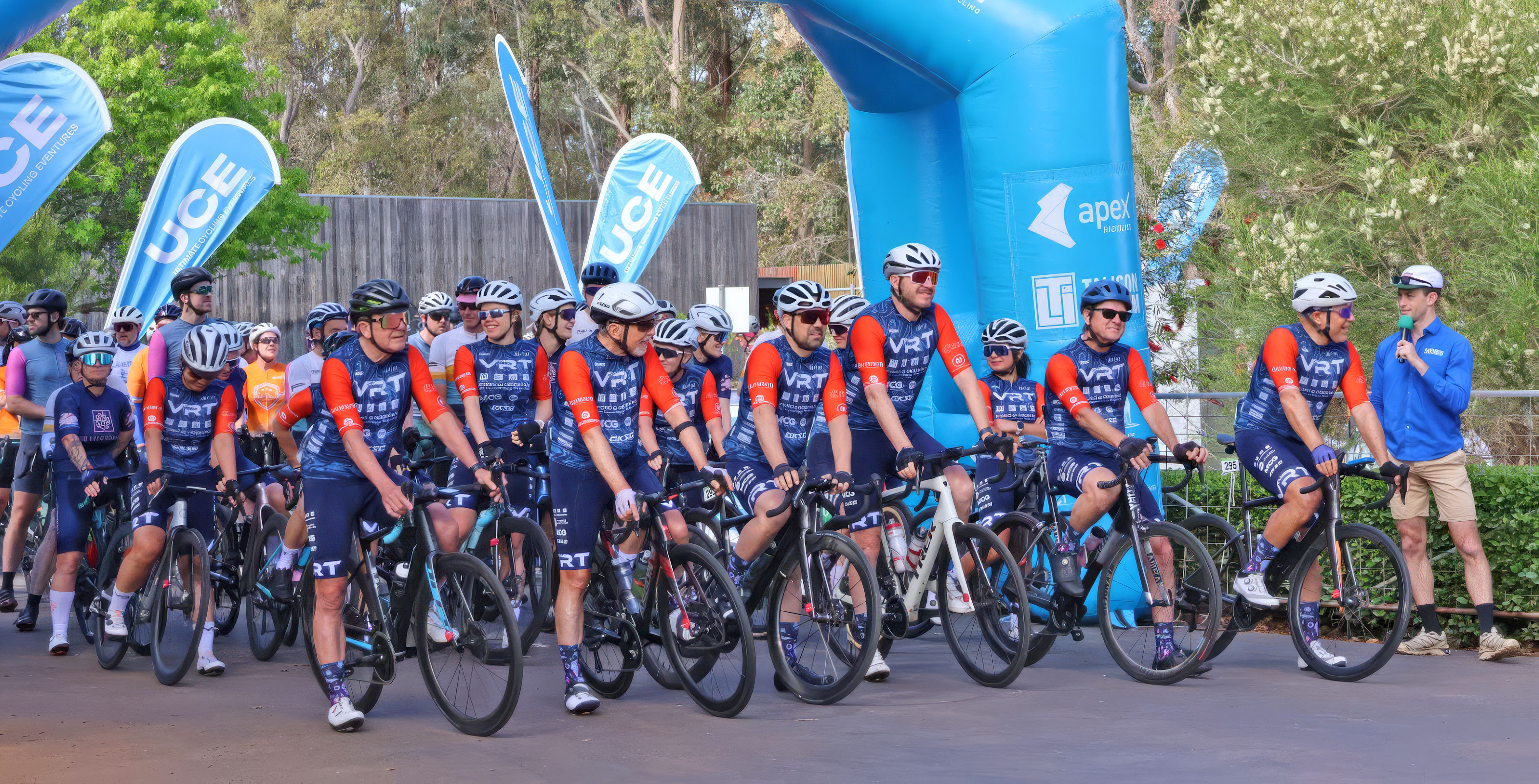 Cycling team in matching blue and orange jerseys on bikes at event start with announcer speaking nearby.