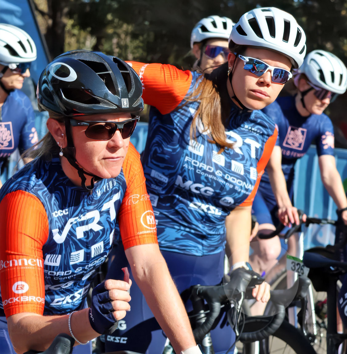 Four cyclists wearing helmets and sunglasses in blue and orange jerseys preparing to race.