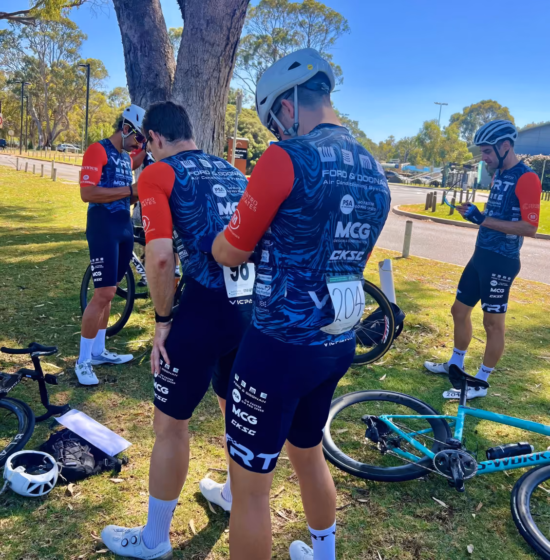 Cyclists in matching blue and red kits prepare by a tree with bikes and gear on grass under clear sky.