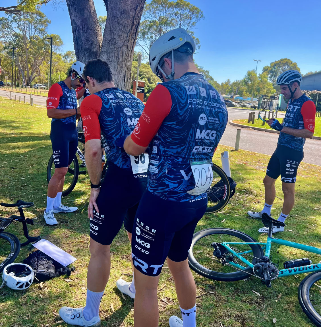 Cyclists in matching blue and red kits prepare by a tree with bikes and gear on grass under clear sky.