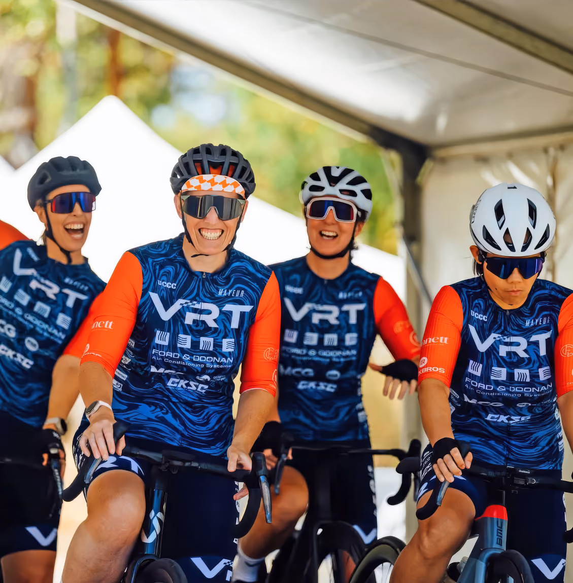 Four cyclists in blue and orange VRT team kits riding indoors, smiling and focused.