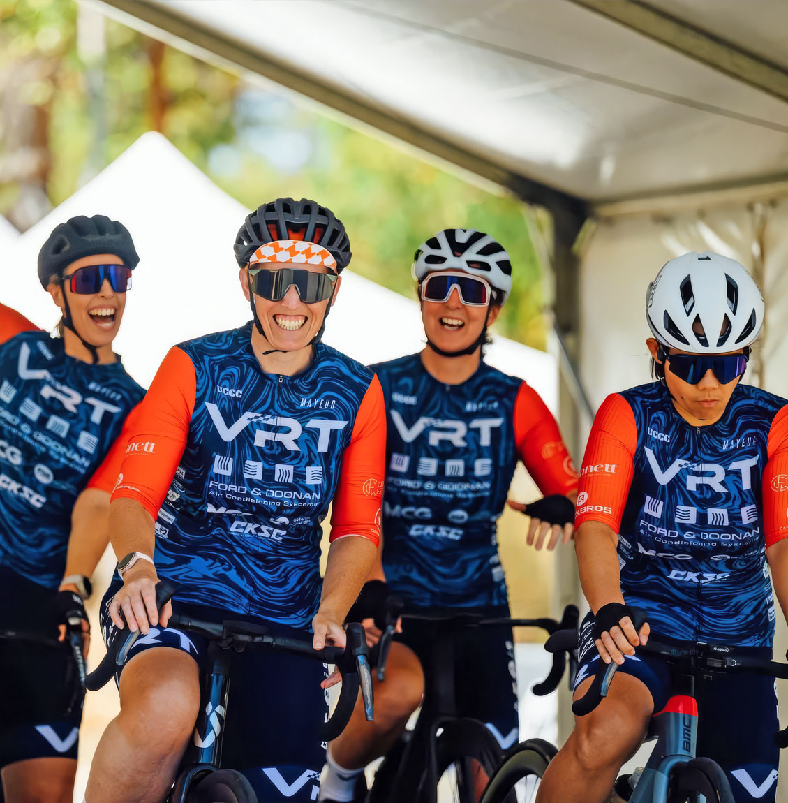 Four cyclists in blue and orange VRT team kits riding indoors, smiling and focused.