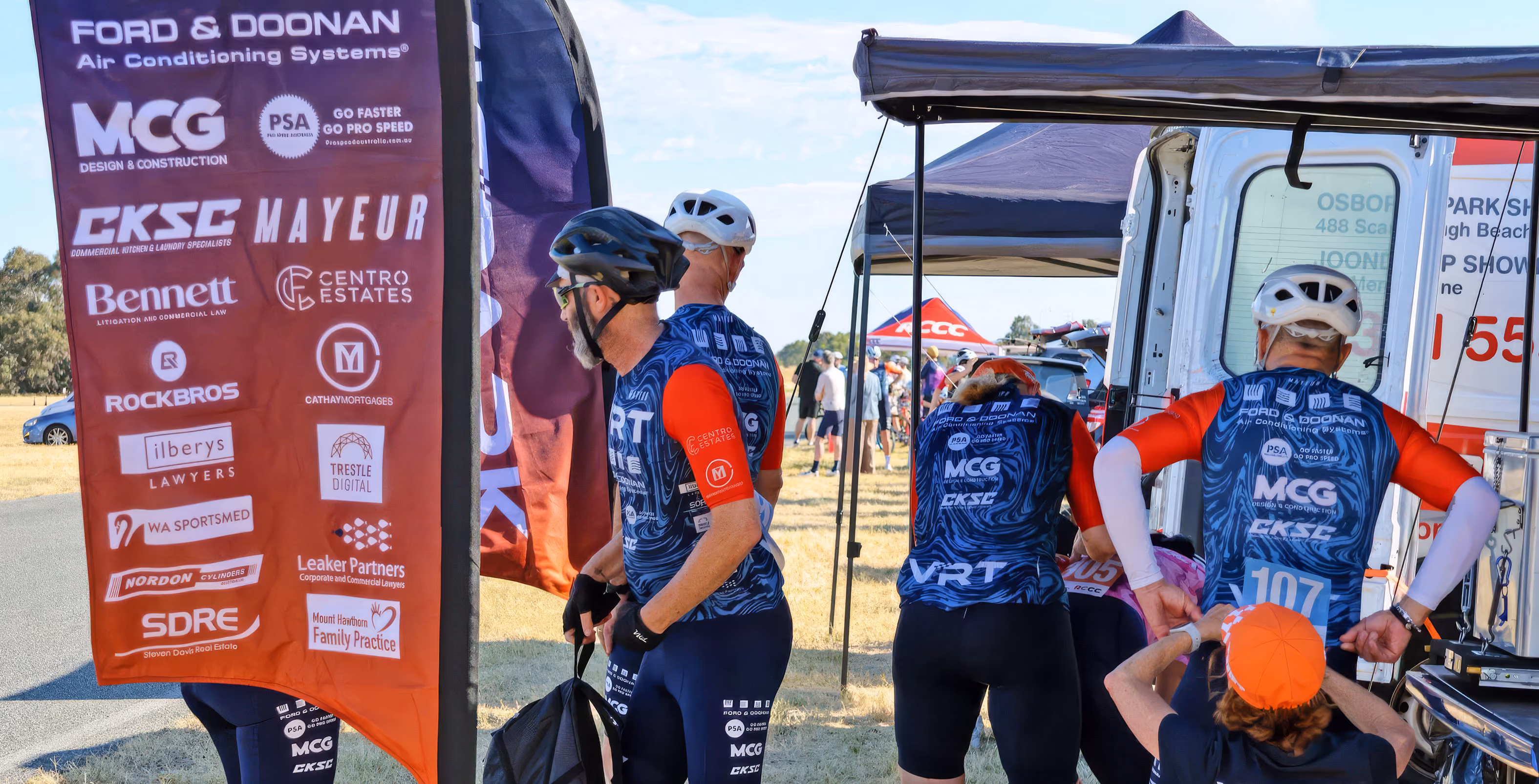 Cyclists wearing blue and red jerseys with sponsor logos preparing near a roadside tent and van.