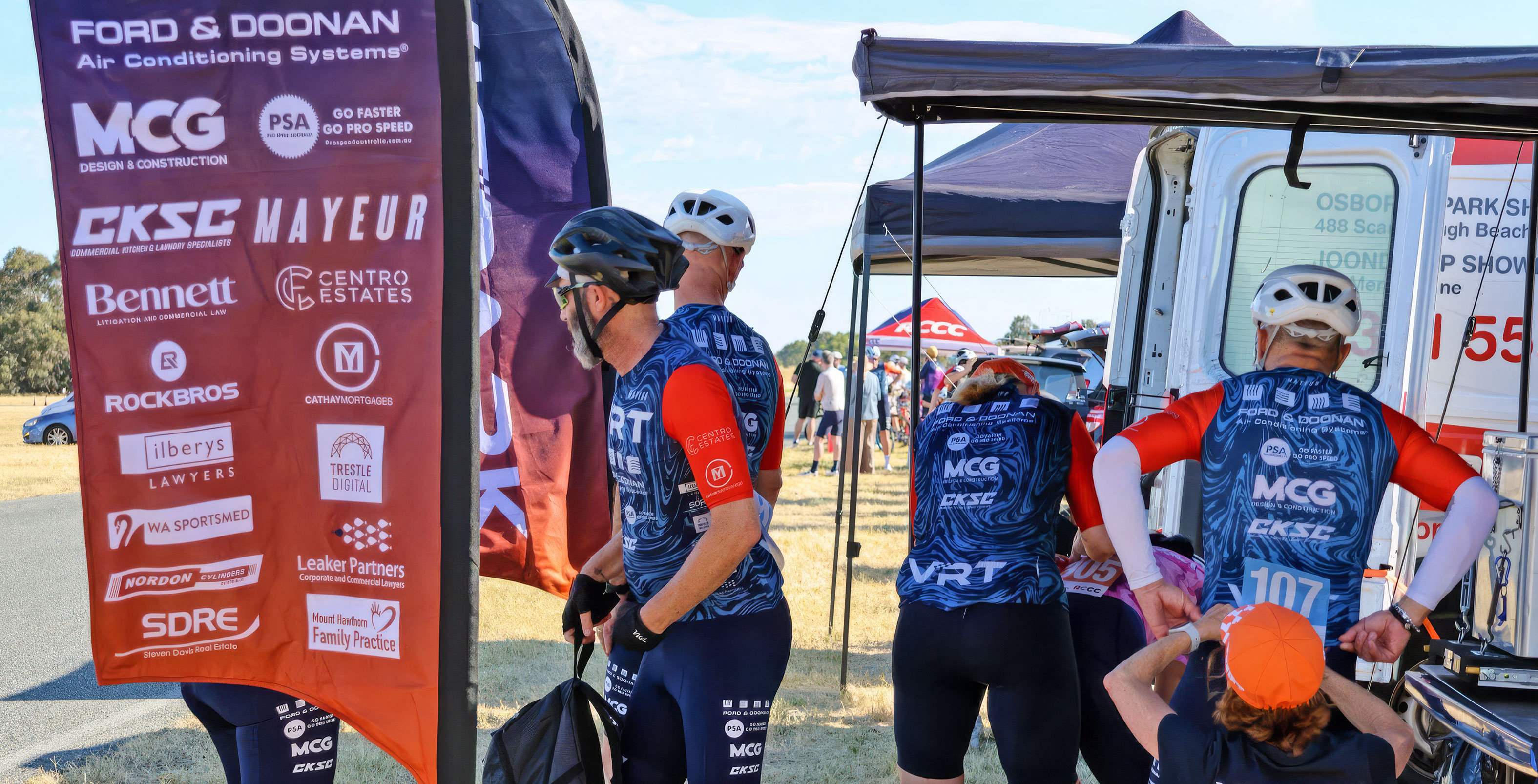 Cyclists wearing blue and red jerseys with sponsor logos preparing near a roadside tent and van.