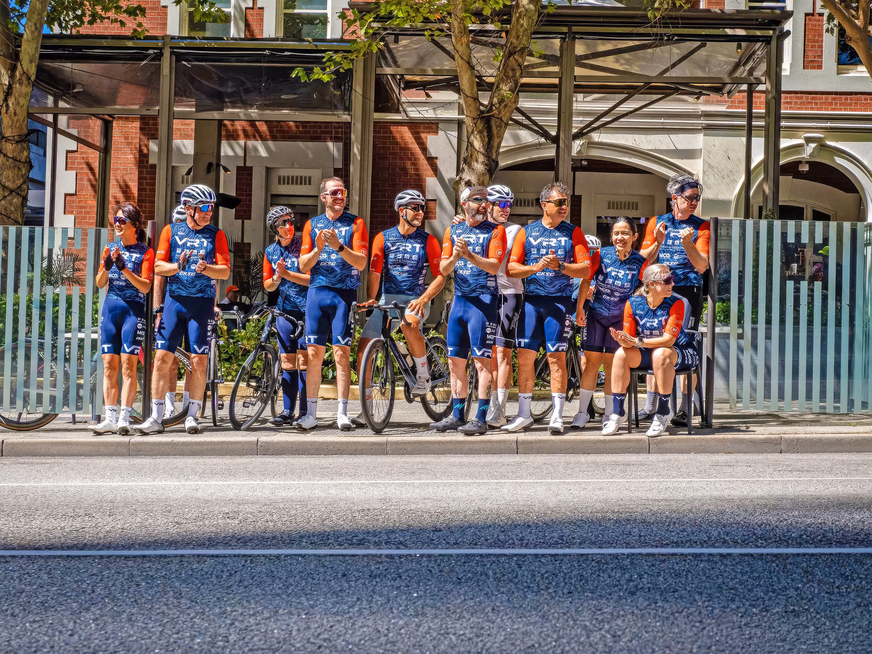 Group of cyclists in matching blue and orange gear standing and sitting at a bus stop on city street.