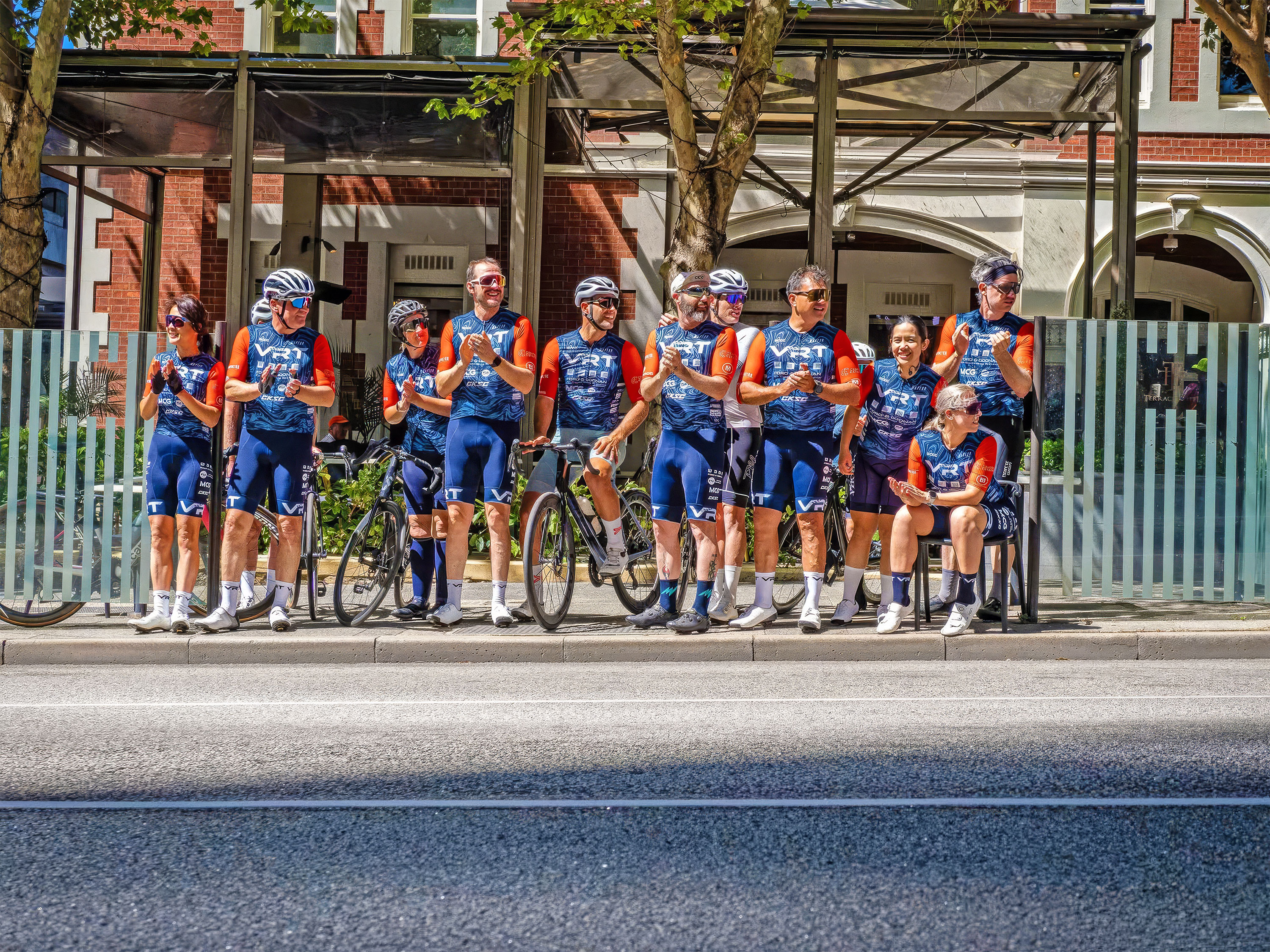 Group of cyclists in matching blue and orange gear standing and sitting at a bus stop on city street.